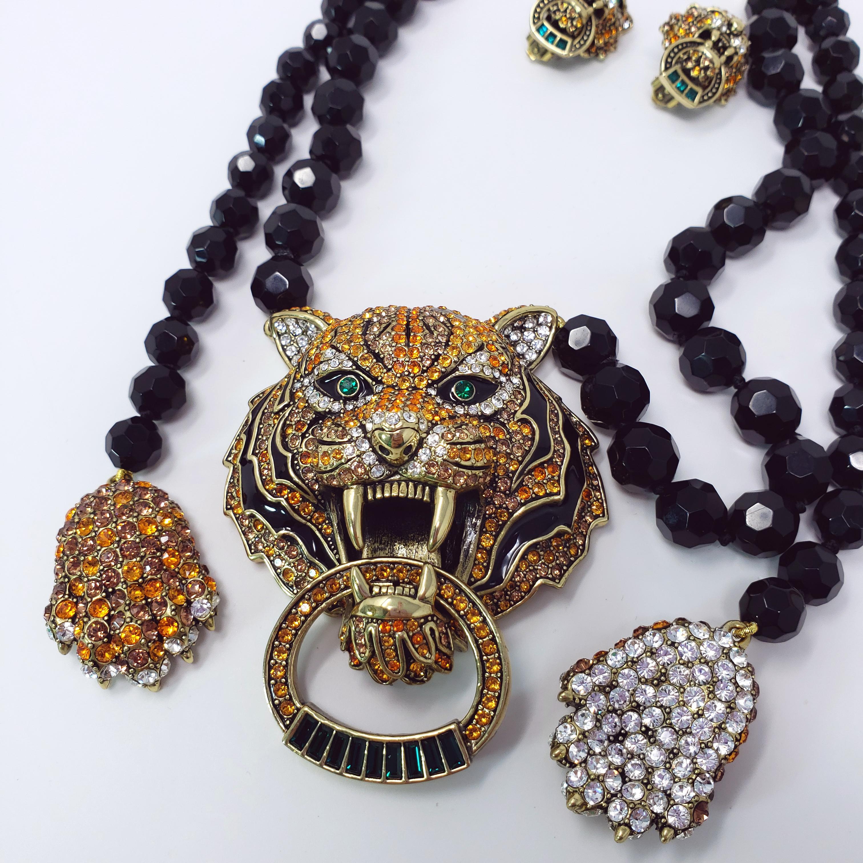 A bold, stunning necklace from Heidi Daus. A fierce tiger decorated with enamel, pave crystals, and two striking green bezel-set eyes holds a doorknocker style ring in its mouth. The tiger head pendant hangs on a single strand of individually