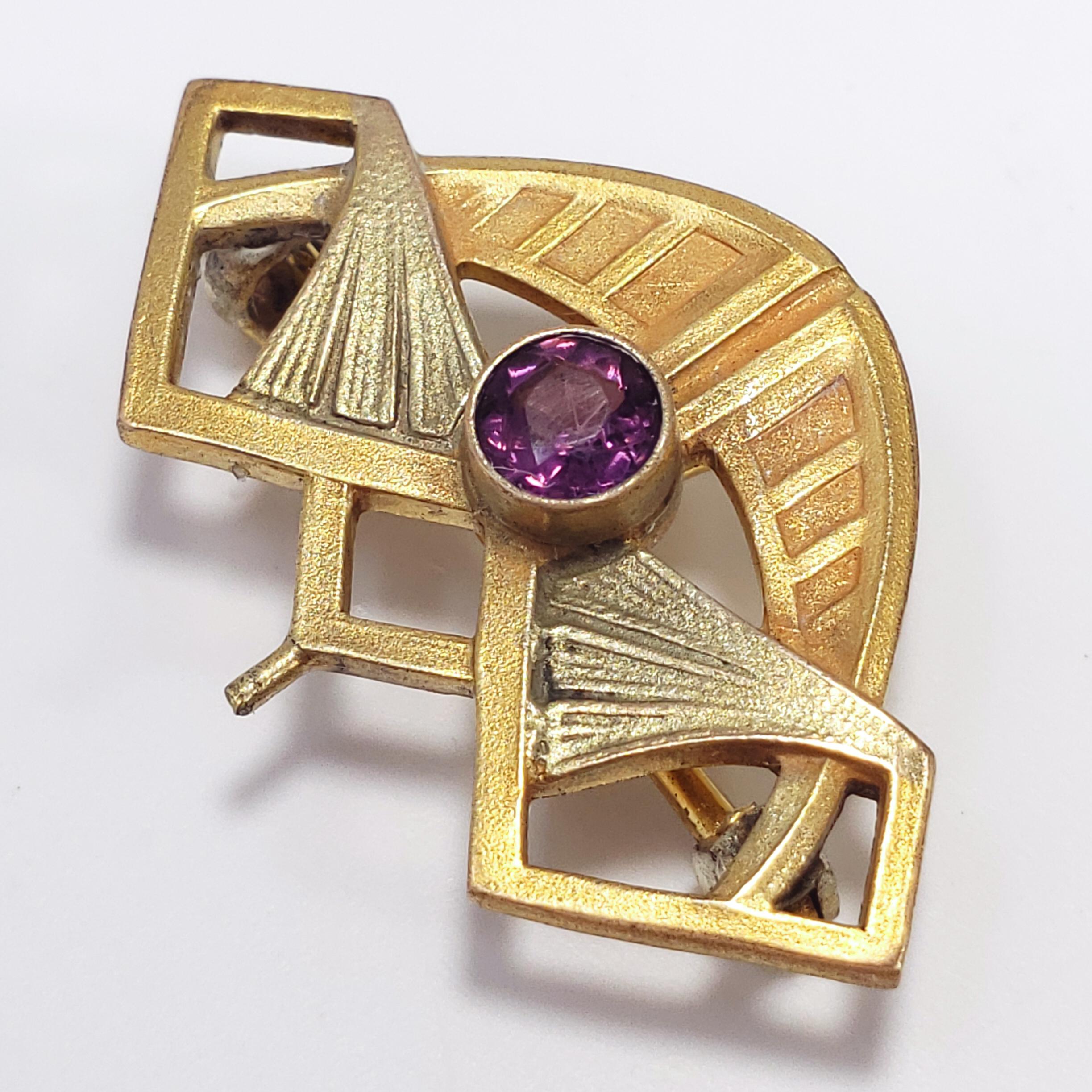 Bold art deco charm on a small accessory for the perfect balance of class and style! Features a single bezel set amethyst-colored crystal in a geometrical brass-tone setting. Can be worn as either a brooch/pin or necklace pendant.

Hallmarks: