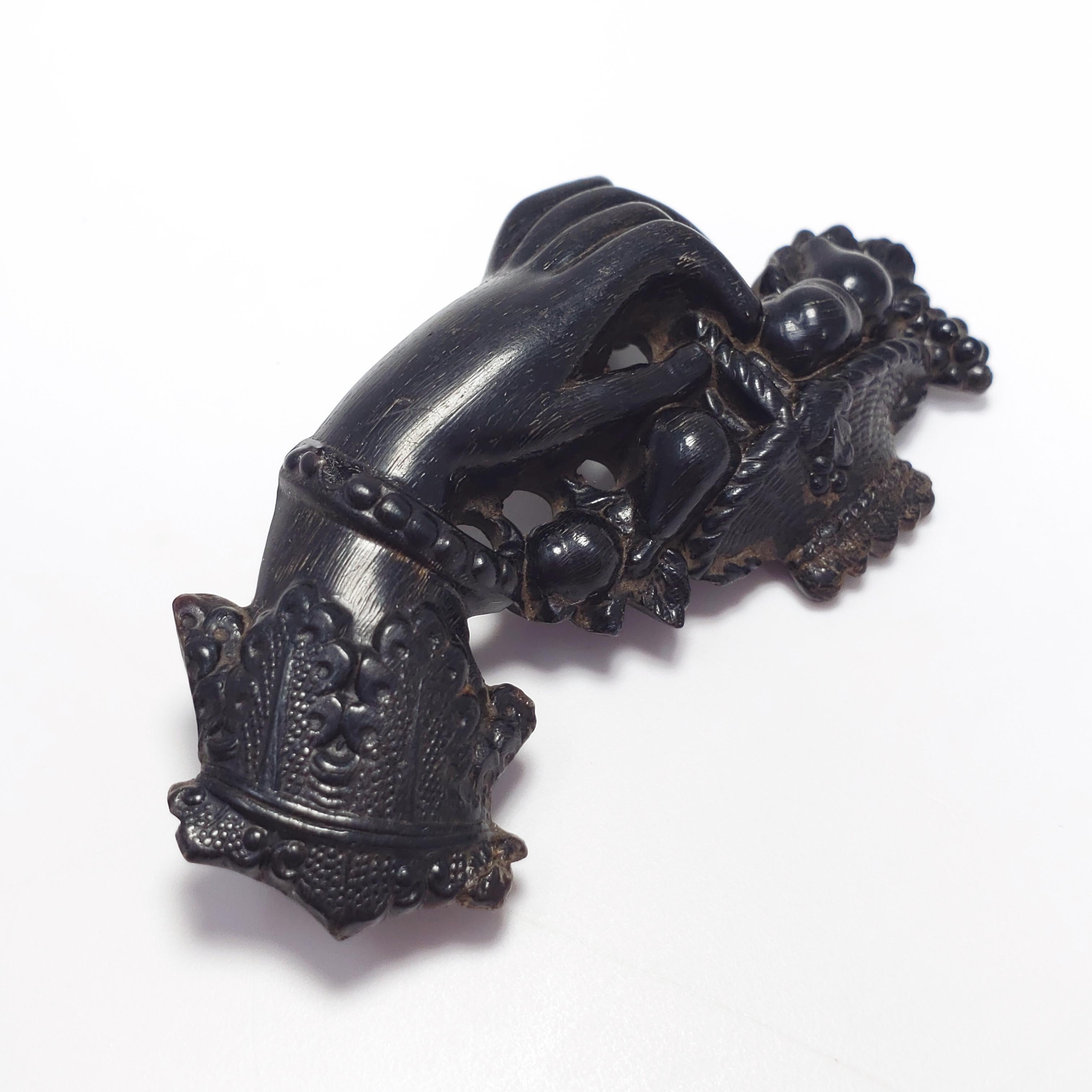 An antique  Victorian pin/brooch made of bog oak, featuring a carved hand & basket motif fastened with brass hardware.