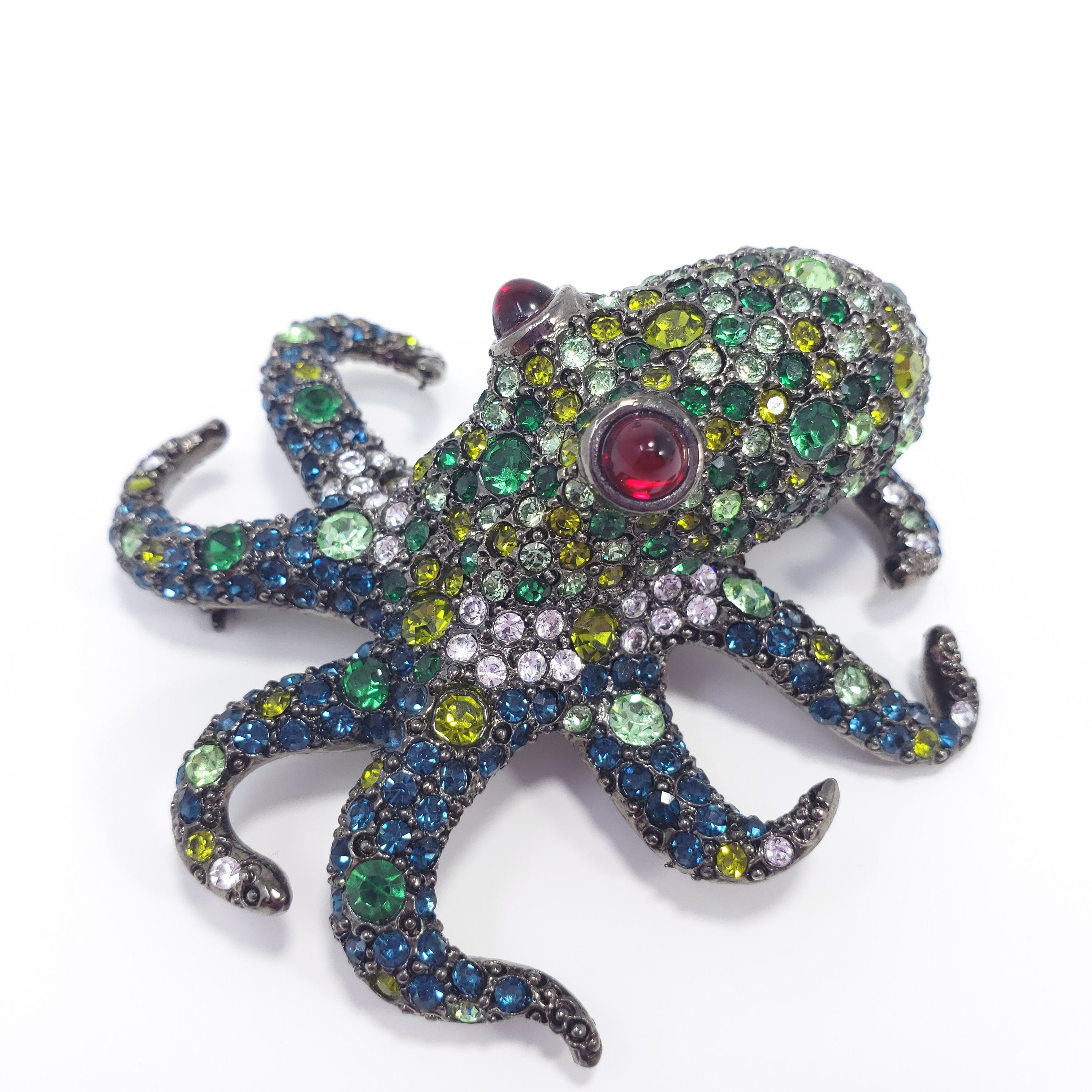 A gunmetal toned octopus decorated with pave crystals in shades of green, accented with two ruby-red cabochon eyes.

By Kenneth Jay Lane.
Hallmarks: Kenneth © Lane