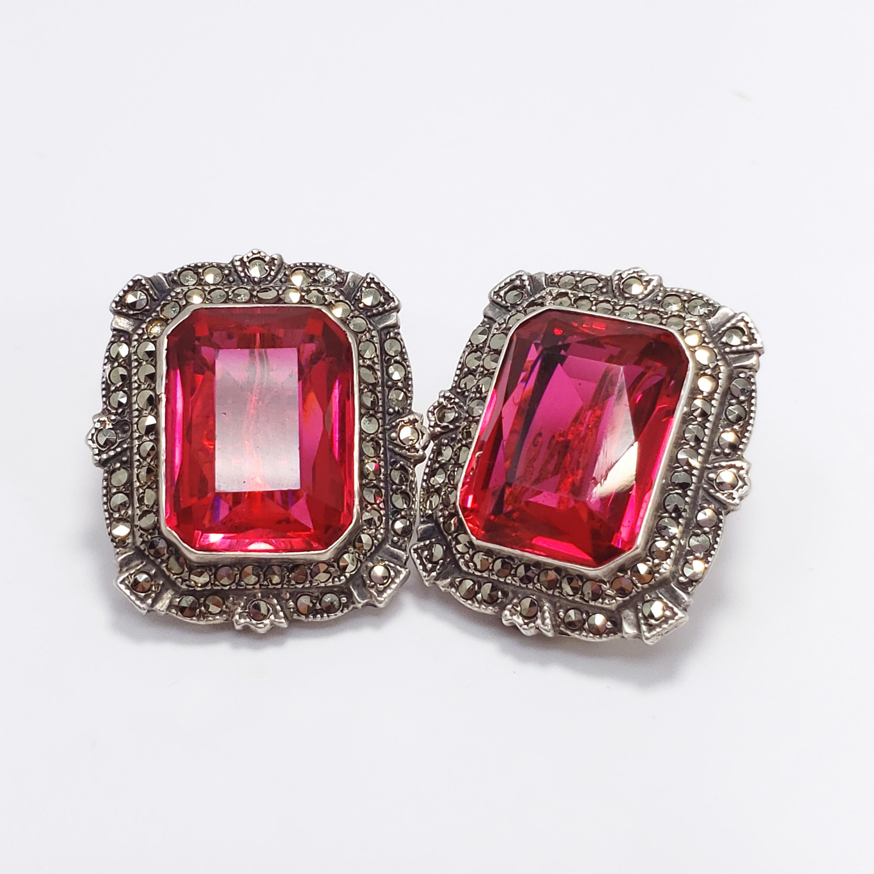 This is a regal-looking pair of earrings from circa late 1880s to early 1900s, with clear Victorian and Art Deco influence. Each earring features a large and bold ruby-colored, open back crystal set in an ornate sterling silver setting. Faceted