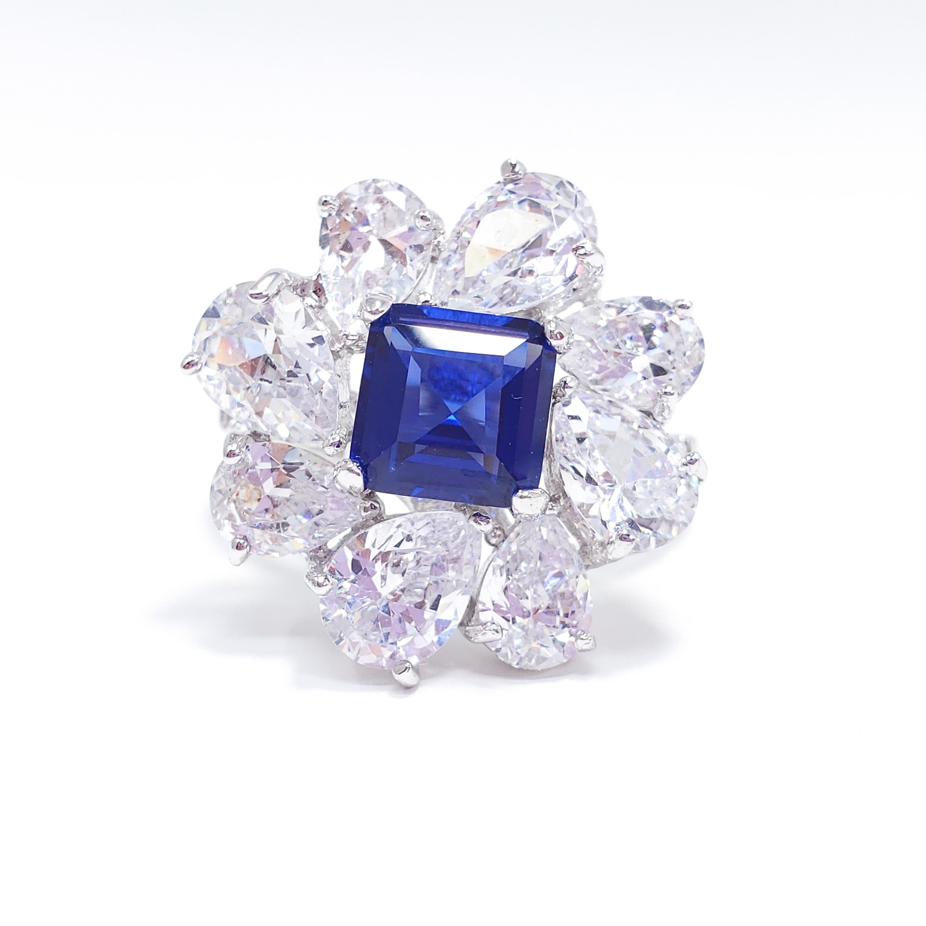 A dazzling cocktail ring by Kenneth Jay Lane. Features a elevated prong-set centerpiece cubic zirconia in sapphire blue, surrounded by clear pear shaped zirconia gems. Smaller crystals accent the front of the band as well. Vibrant rhodium plated