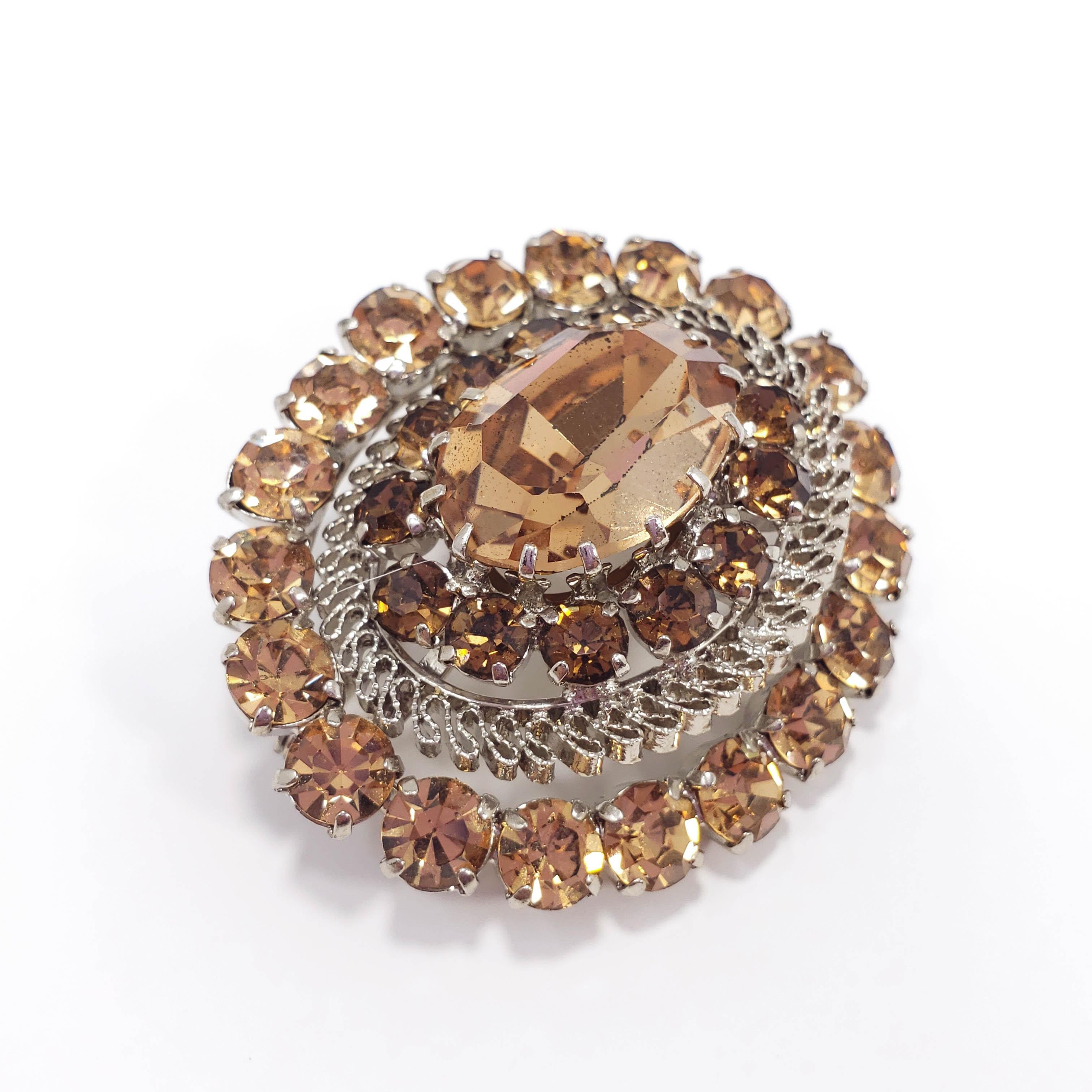 An elegant pin featuring a large centerpiece topaz-colored, open foiled-back crystal, prong-set in a silvertone setting and surrounding with smaller prong-set crystals and filigree accents. The center setting is angled back relative to the outer