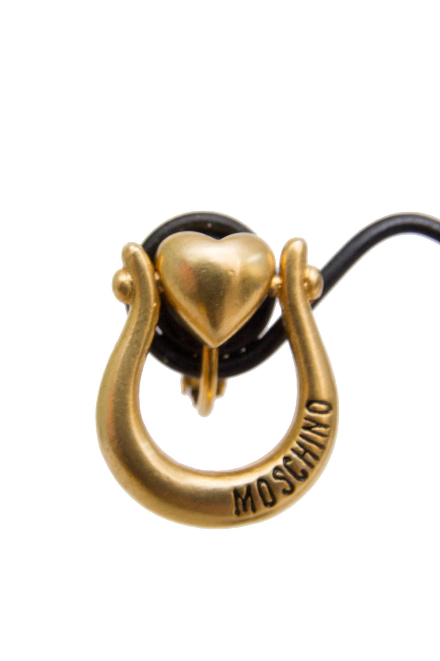 A fun pair of 1990s Moschino clip on earrings that consist of a delicate heart surrounded by what appears to be a small horseshoe. Both earrings are stamped with 'Moschino' in black lettering on the front. 

