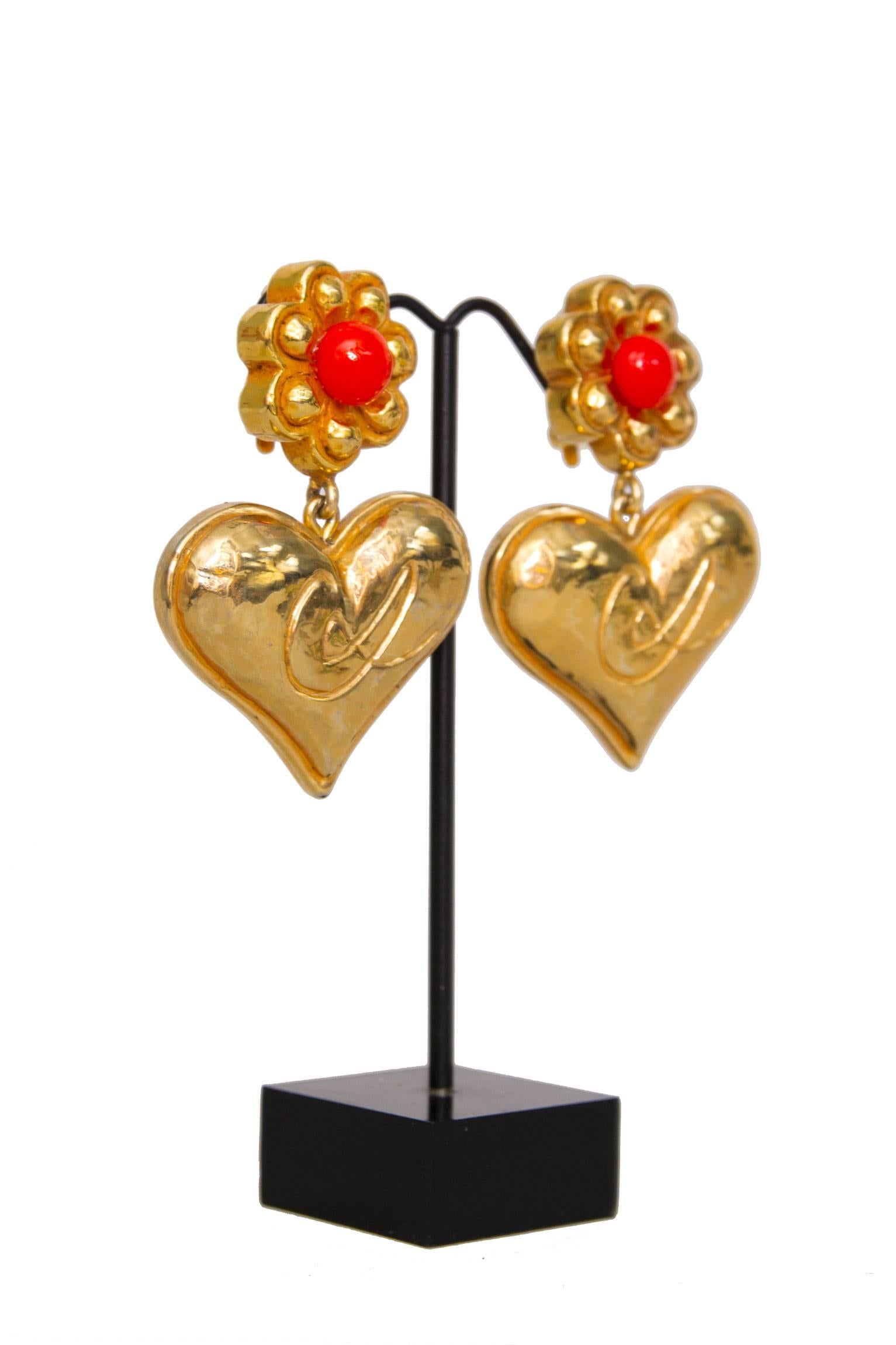 A pair of 1980s Christian Lacroix goldtone clip-on earrings with a heart pendant. At the top the earrings have a small flower which is set with a bright orange stone. The heart shaped pendent is attached by a small chain link and  written across is