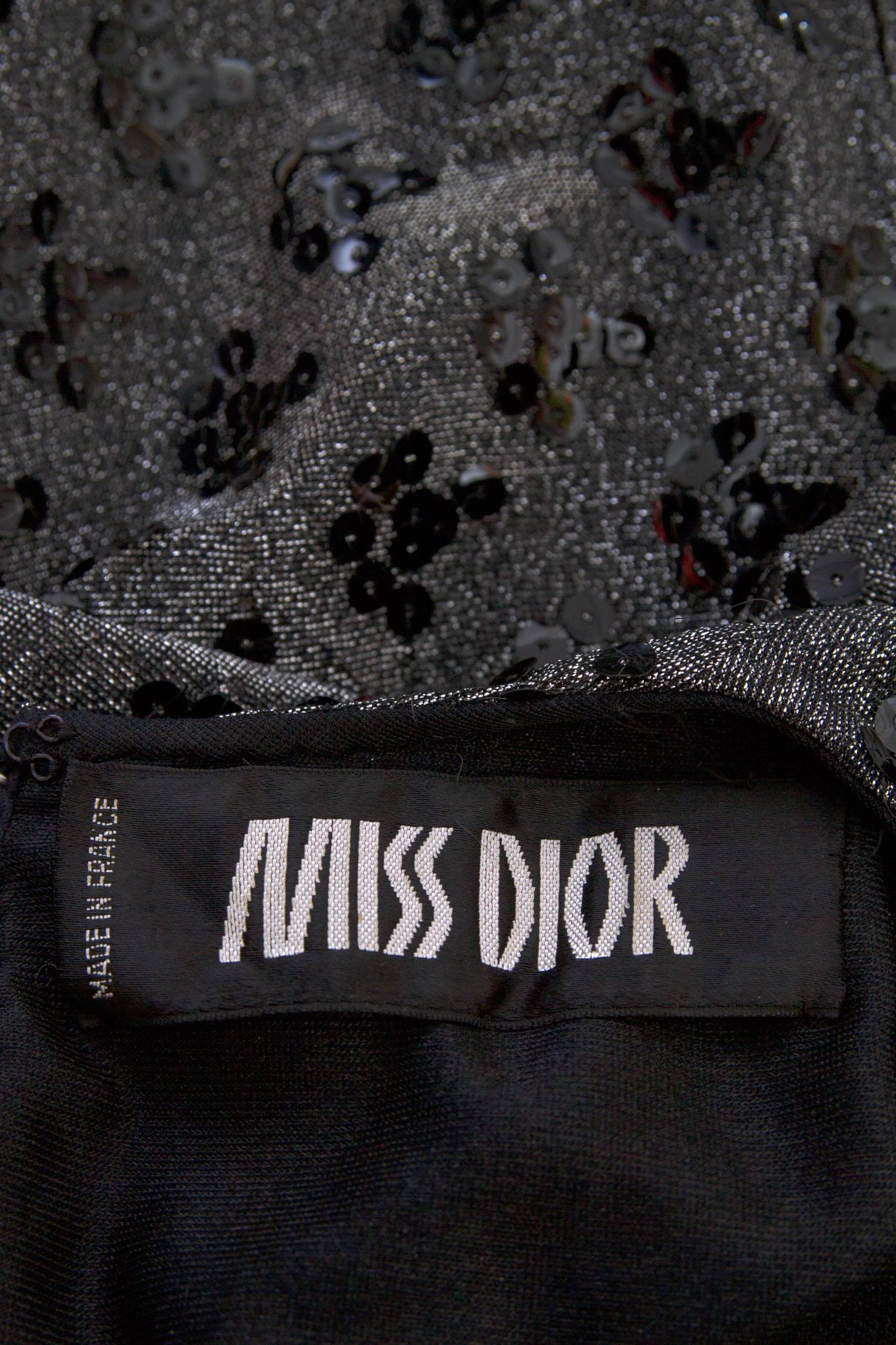 A glamorous 1960s silver lurex cocktail dress by Miss Dior. The dress has a drop waistline, a round neckline and long tapered sleeves. The pleated skirt is separated fromt the fitted bodice by a black beaded trim. The dress is embellished with small