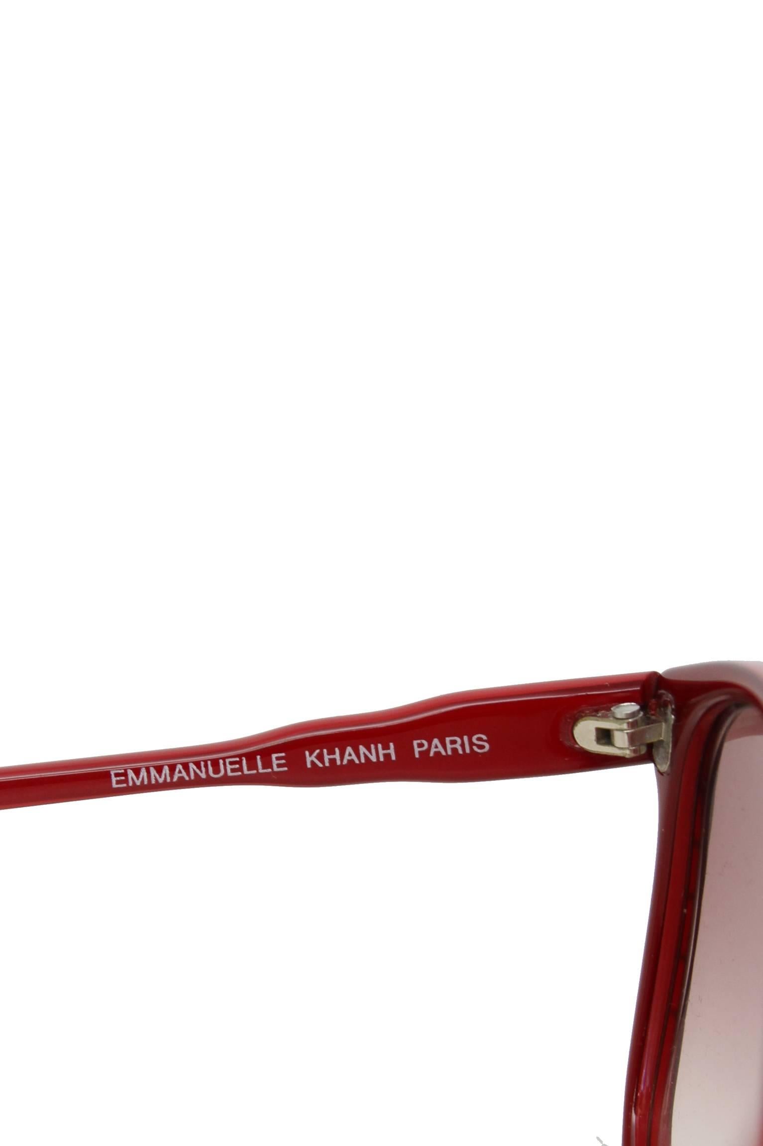 A pair of 1970s Emmanuelle Kahn red  frame sunglasses with a small gold colored logo situated at the top of each temple, and the red lenses are situated in a square frame with gold details in the outer top corners. 

The logo measures: 0.8x 0.8