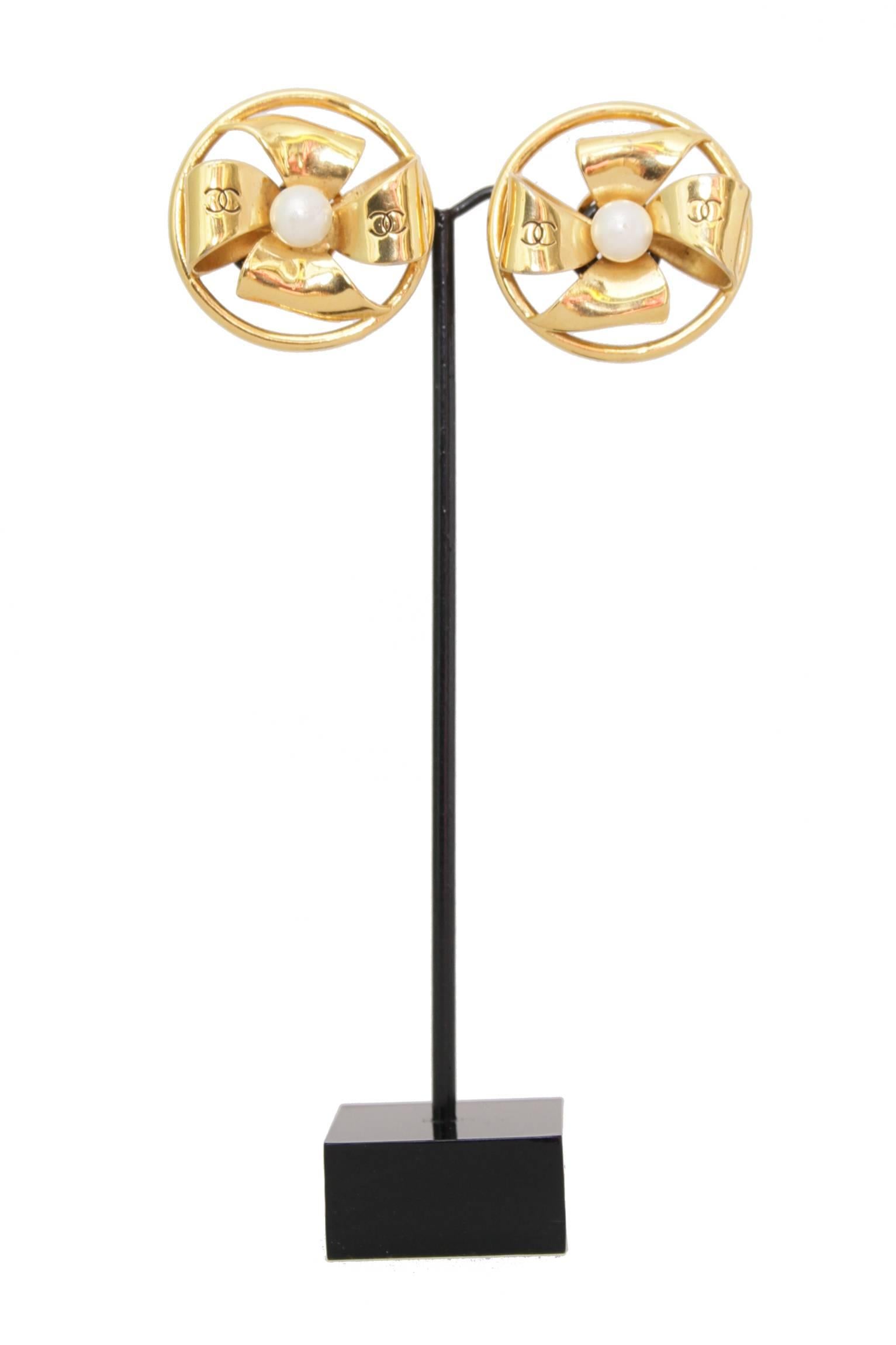 A pair of 1980s large gold toned Chanel clip-on earrings shaped as a bow set in a round gold wire. A Chanel double 'C' logo is engraved on each side of the bow encasing a mother of pearl center. 

The earrings are stamped: Chanel. 