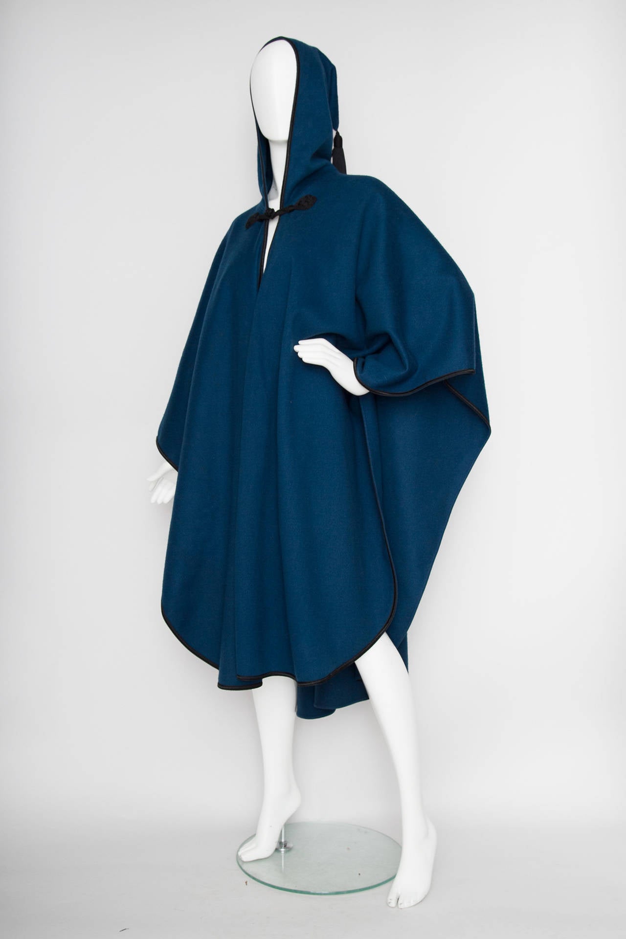 A rare 1970s Yves Saint Laurent unlined teal blue wool cape with black silk tapestry trim, an oversized hood with a centered black tassel. 

The cape was made for 1976 Fall/Winter Yves Saint Laurent Rive Gauche  