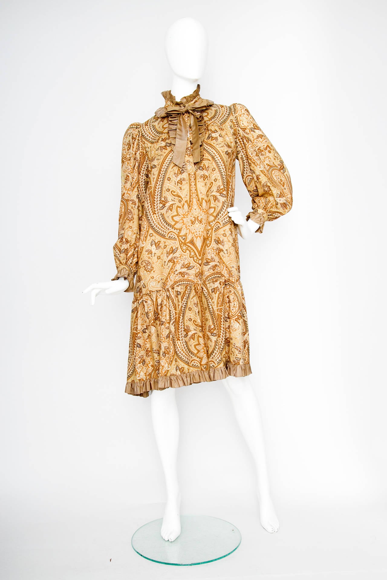 A stunning 1970s two piece Yves Saint Laurent ensemble consisting of an a-line peasant dress with an allover paisley print in a beige and light brown color scheme and a cropped quilted jacket. 

The dress features long sleeves, light brown ruffle