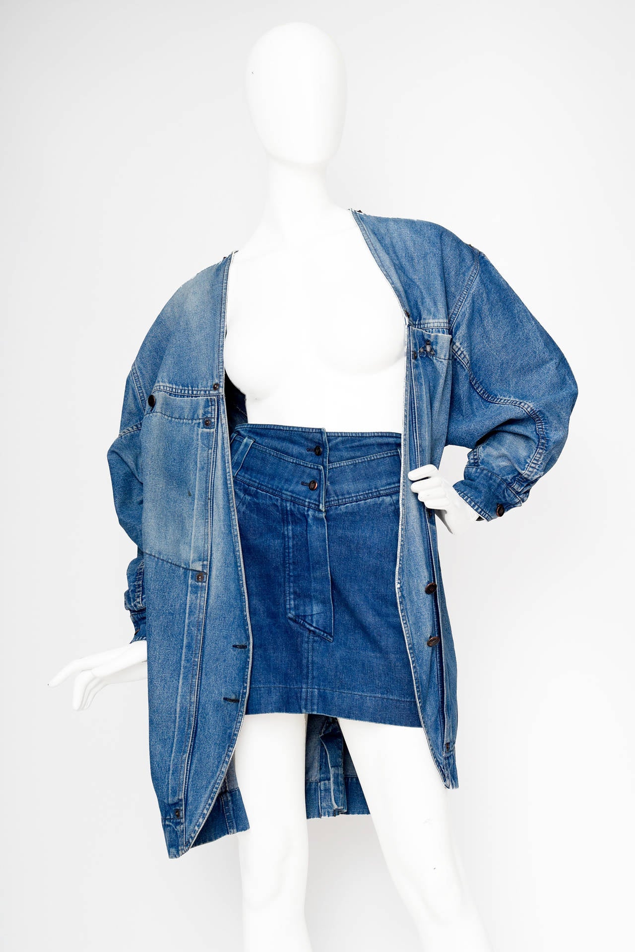 A 1980s Claude Montana denim jacket and mini skirt ensemble. The jacket has an oversized fit and has two exaggerated patch pockets on the bust and two side pockets. The jacket has a deep v-neckline and closes in the front with a two button closure.