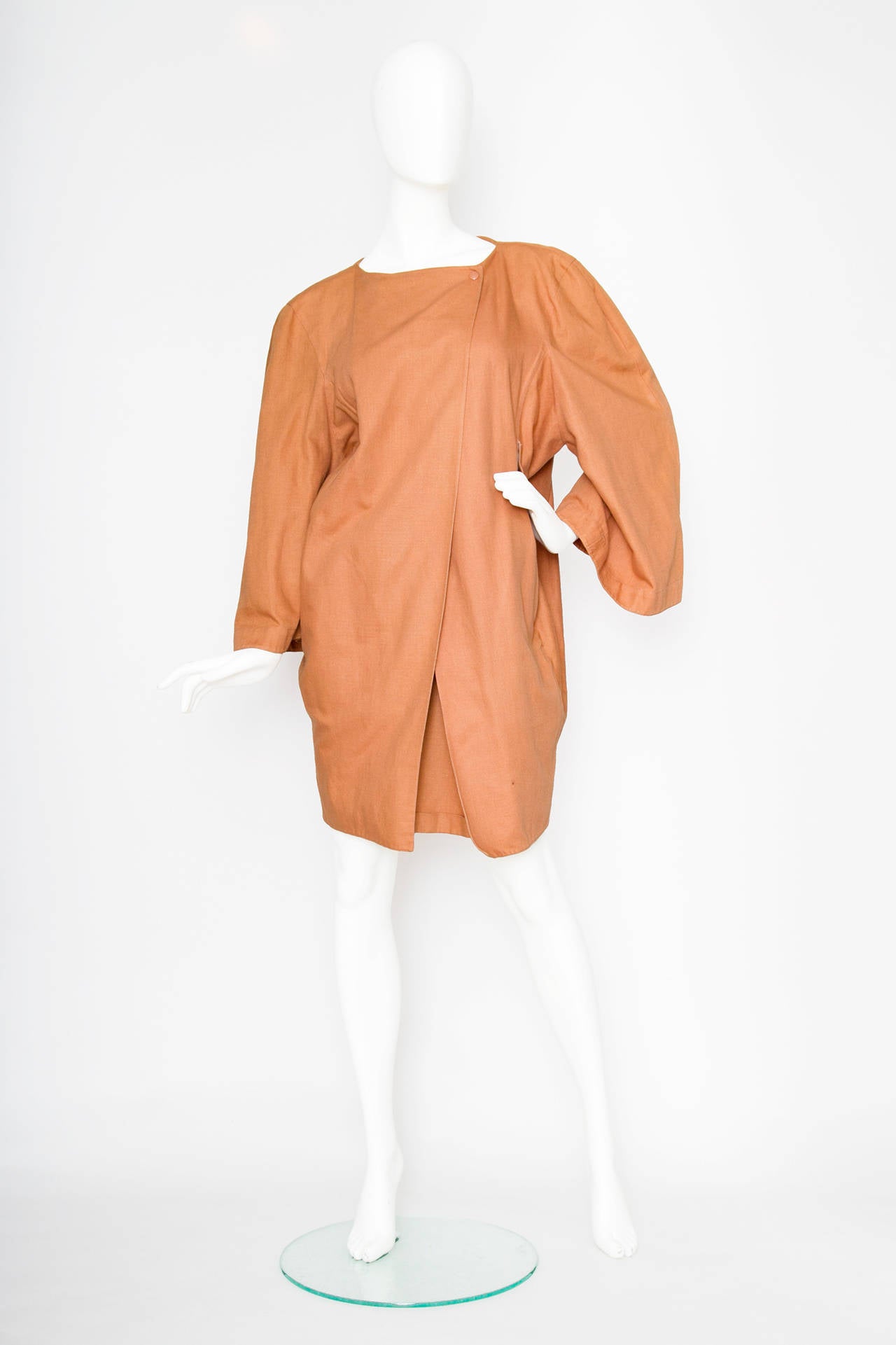 A 1980s Claude Montana orange linen jacket and short ensemble. The jacket has an oversized fit and rounded shoulders with wide sleeves. The jacket has a round neckline and closes in the front with a cross body closure fastened with a push button and