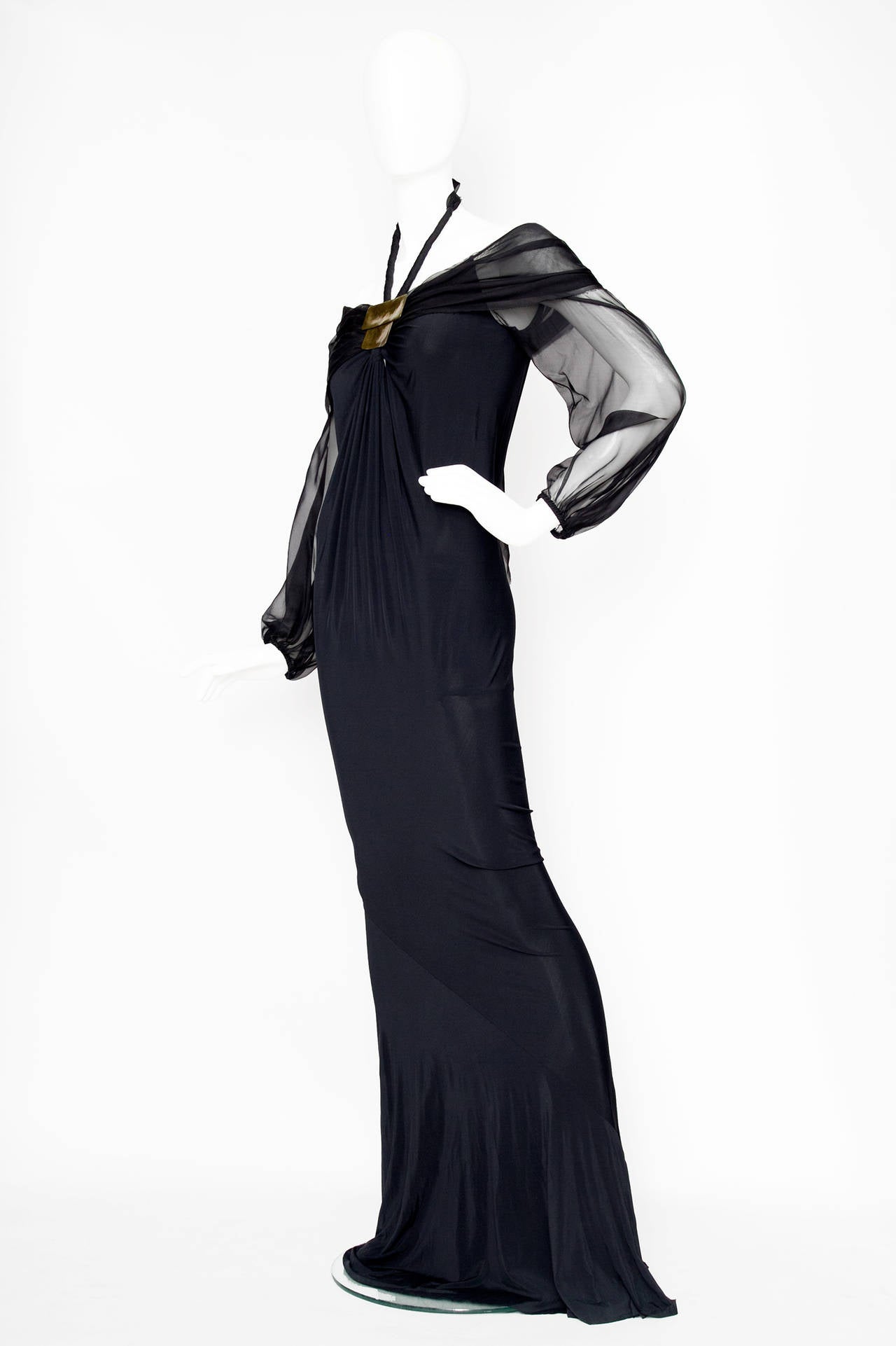 A stunning 1990s Donna Karan floor-length black evening gown in silk jersey. The dress has long sheer sleeves and an off the shoulder neckline, both in a light silk chiffon. In the front the two fabrics are gathered by a gold metal plate detail and