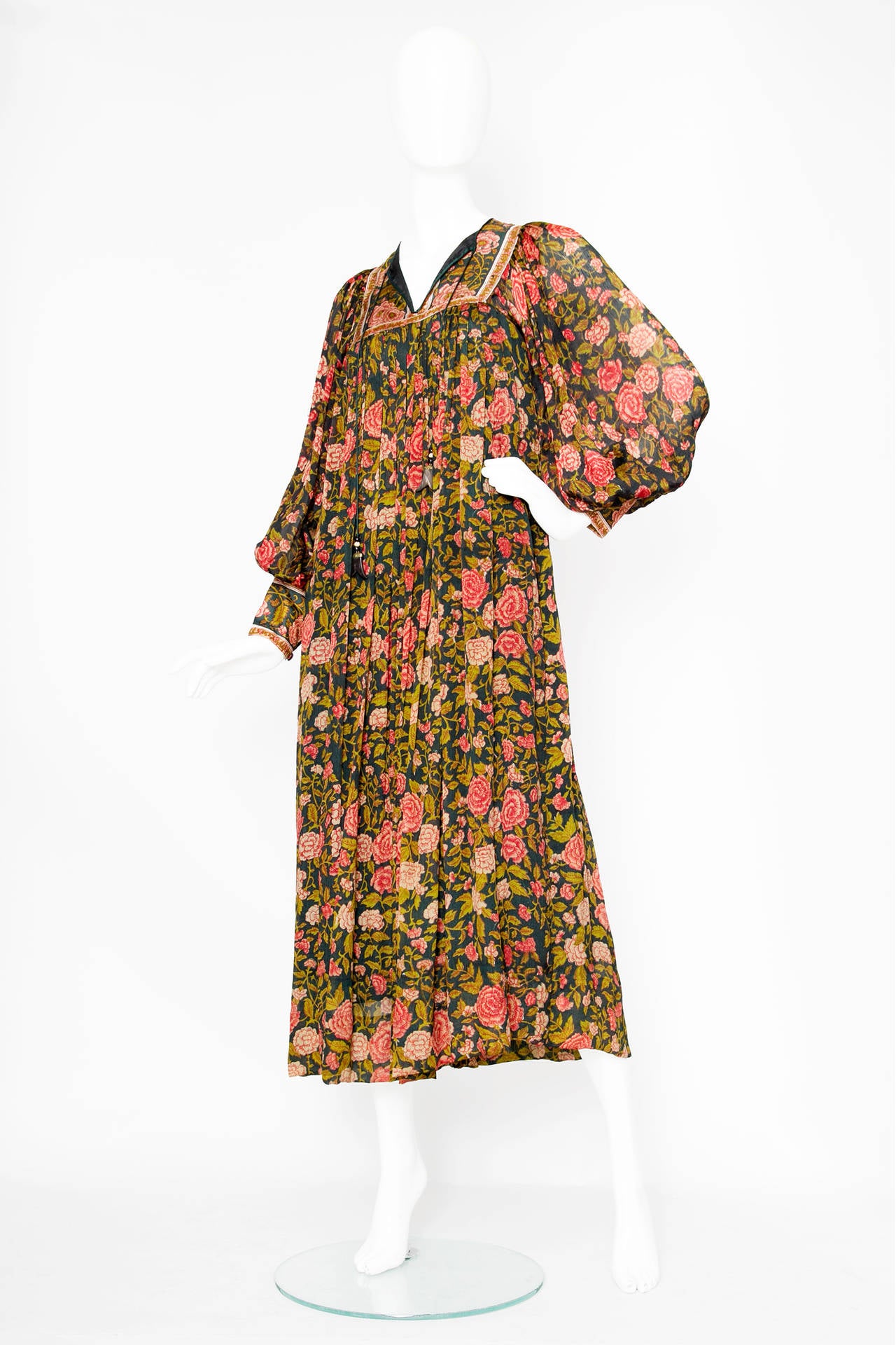 A 1970s hand block printed silk kaftan with an all over flower print in deep red and green hues that gently flows across the garment. The kaftan has sheer voluminous sleeves with three buttoned cuffs. The Dress is fully lined and have a tie string