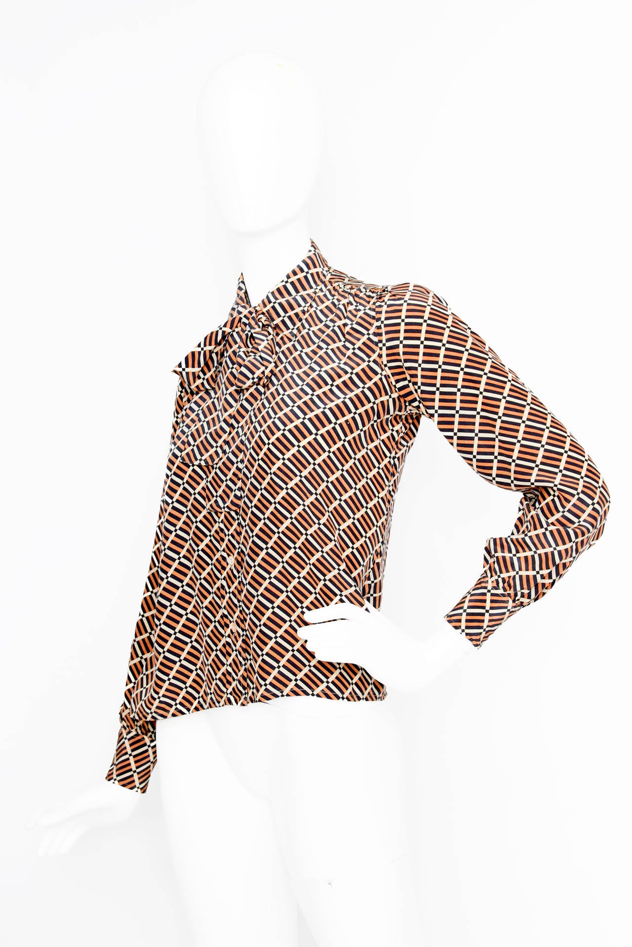 A 1970s Yves Saint Laurent silk shirt with an all over graphic print in brown, black and white. The shirt has long sleeves with two buttoned cuffs and a tie ribbon detail attached under the collar. The shirt closes down the front with pale orange