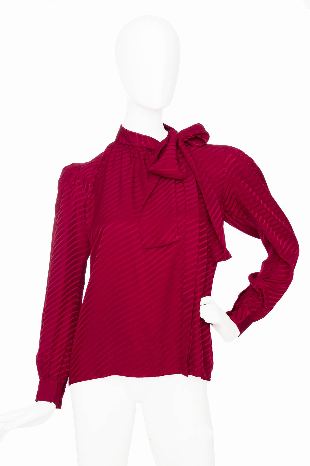 A 1980s Yves Saint Laurent jacquard silk blouse with an asymmetrical and invisible front button closure. The shirt has one buttoned cuffs and and attached  bow to tie around the neck. The shoulders are slightly puffed and tulle shoulder pads.