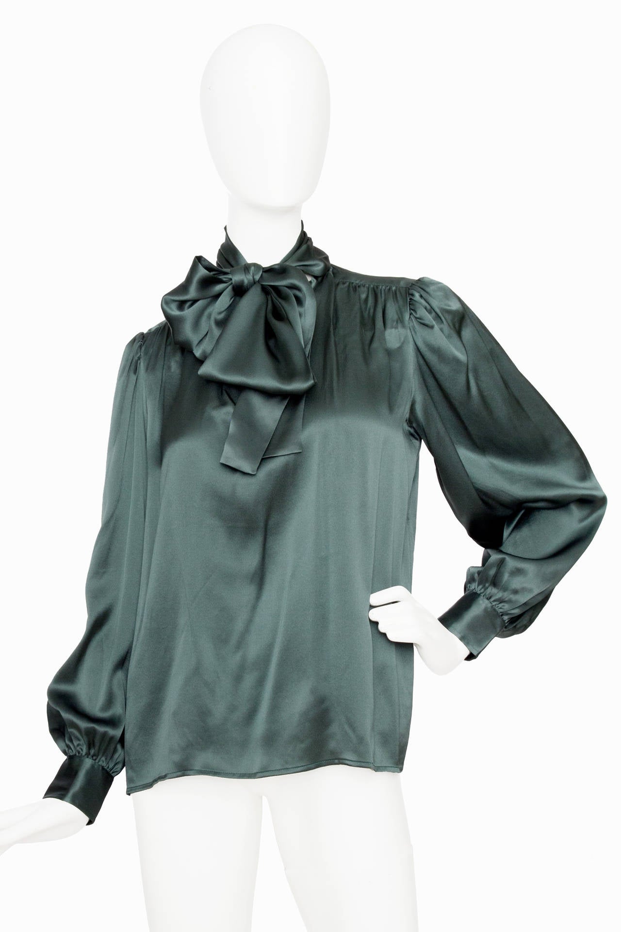 A 1980s Yves Saint Laurent dusty green silk blouse with blouson sleeves and slightly puffed shoulders. In the front the blouse has a single one button closure and an attached bow to tie around the neck. The blouse has one buttoned cuffs. 

The