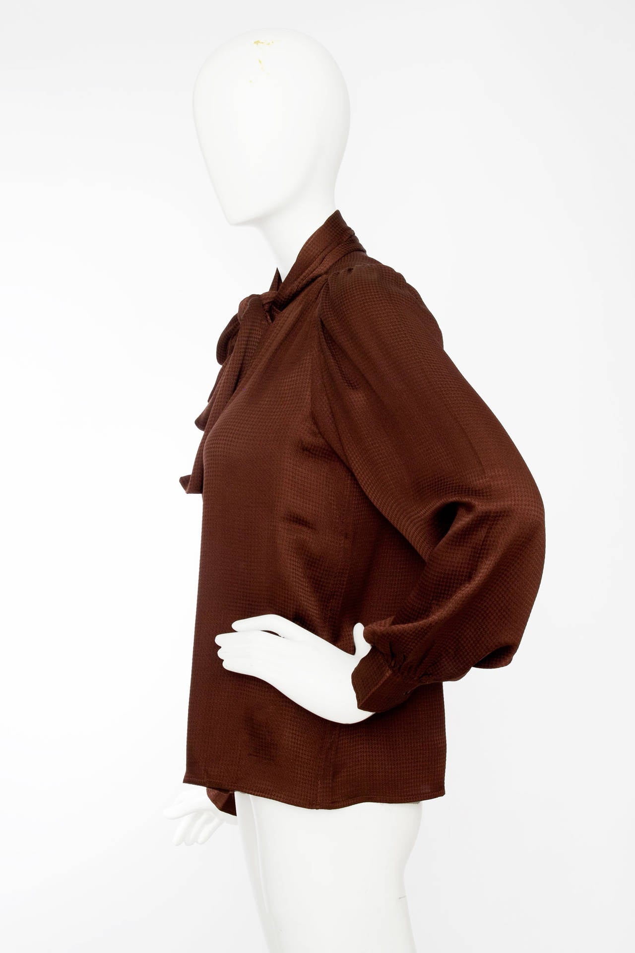 A 1980s Yves Saint Laurent chocolate brown silk blouse with a subtle houndstooth jacquard weaving and an attached scarf around the neckline. The blouse has a split front with a single button closure and one buttoned cuffs. 

The size of the blouse