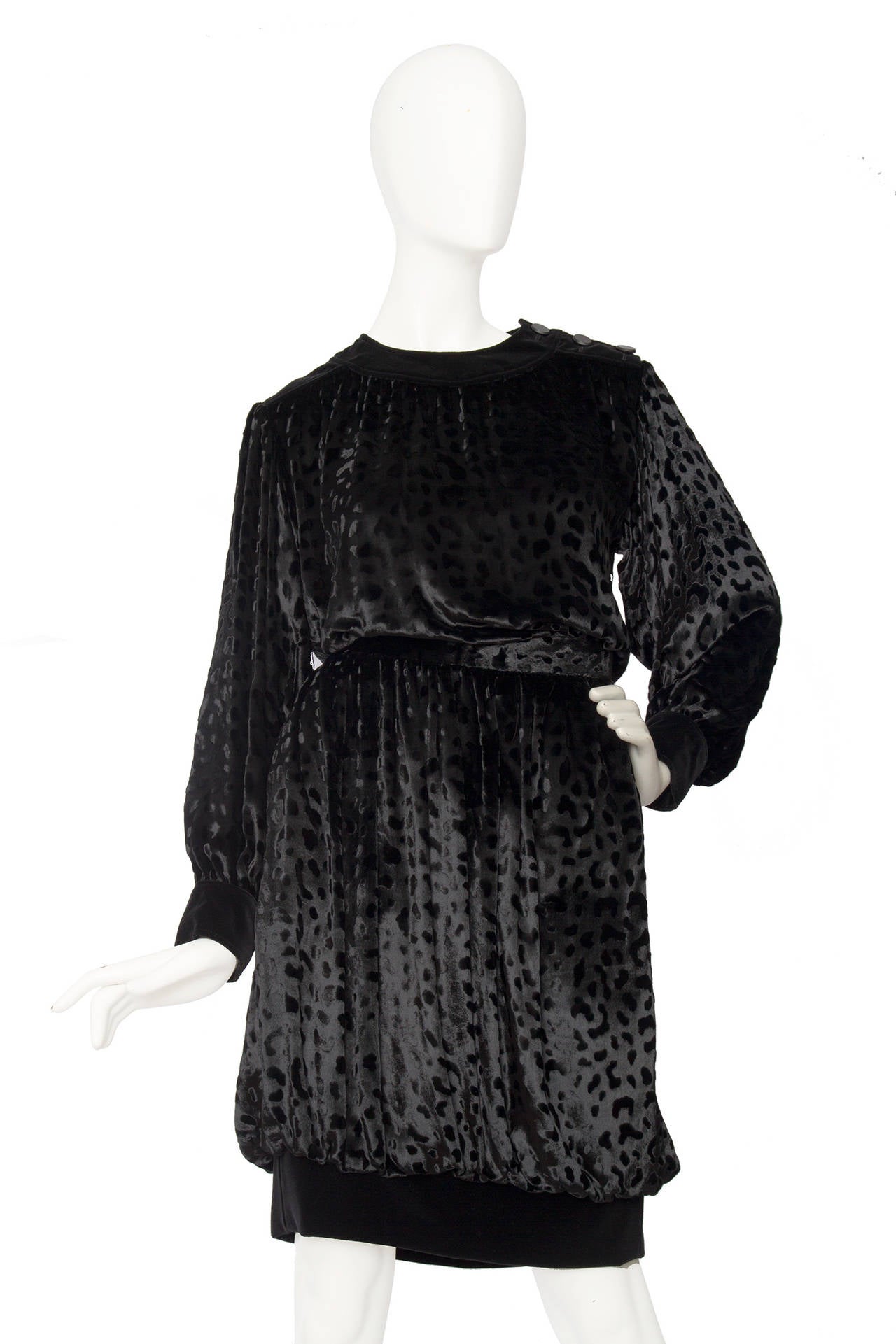 A 1980s Yves Saint Laurent two-piece black silk velvet ensemble consisting of a loosely fitted blouse and a balloon skirt. 
The blouse has a wide velvet trim around each cuff and around the neckline, which is completed by a three-buttoned opening