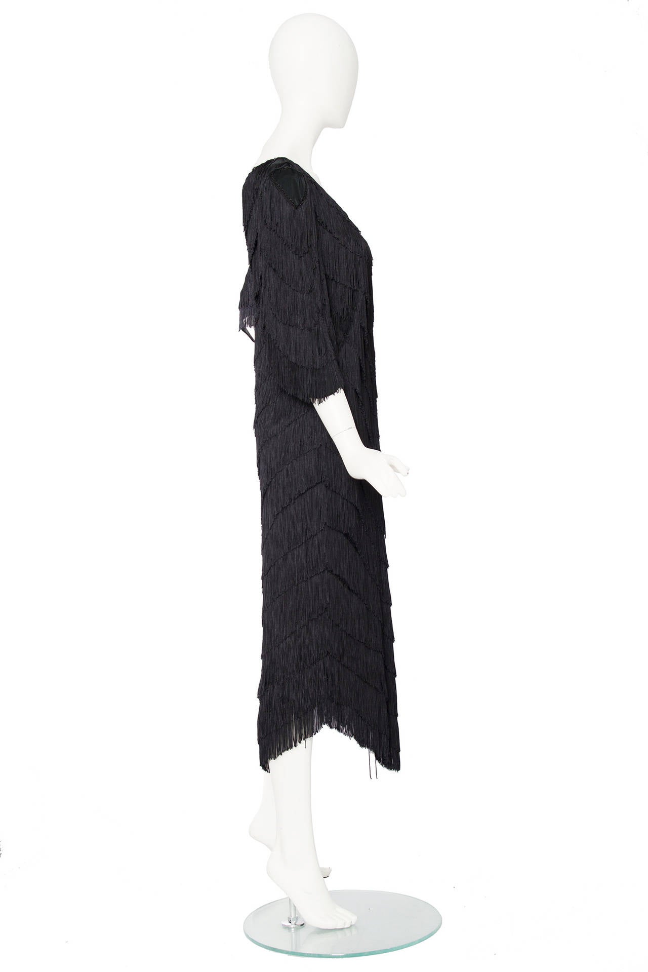A 1980s Norma Kamali black fringe dress with an asymmetrical hemline and fringe detailing. The neckline and hem ascends into a triangular shape matching the fringe. The three quarter length sleeves have slight volume and the shoulders have small