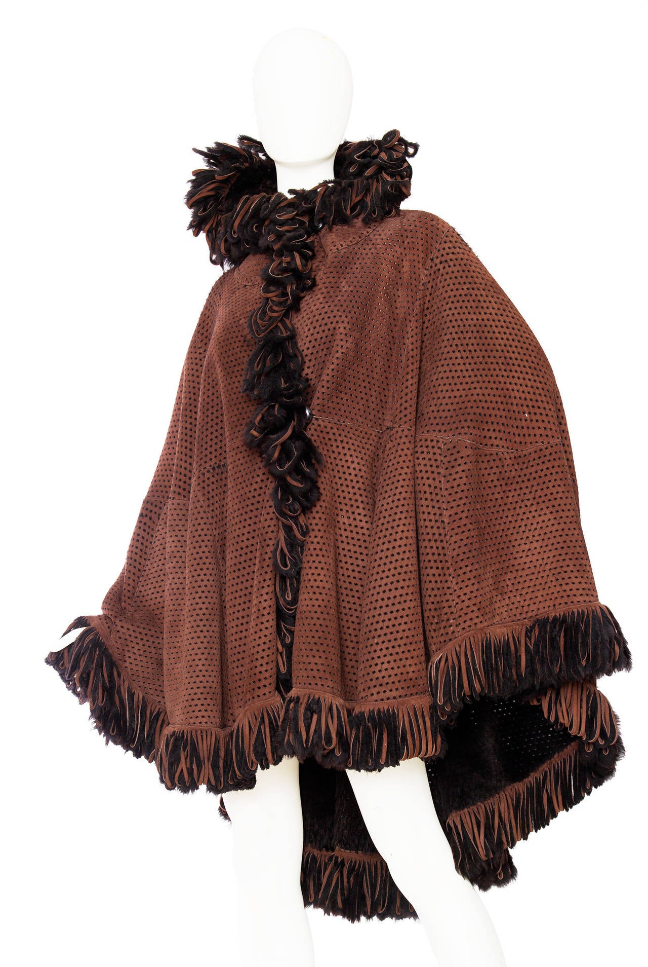 A 1980s brown and black Fendi perforated shearling hooded cape with fringe trim detailing around the hemline and down the front. The cape has front button closure on the top and has a round hemline that falls lower on the back. 

The size of the