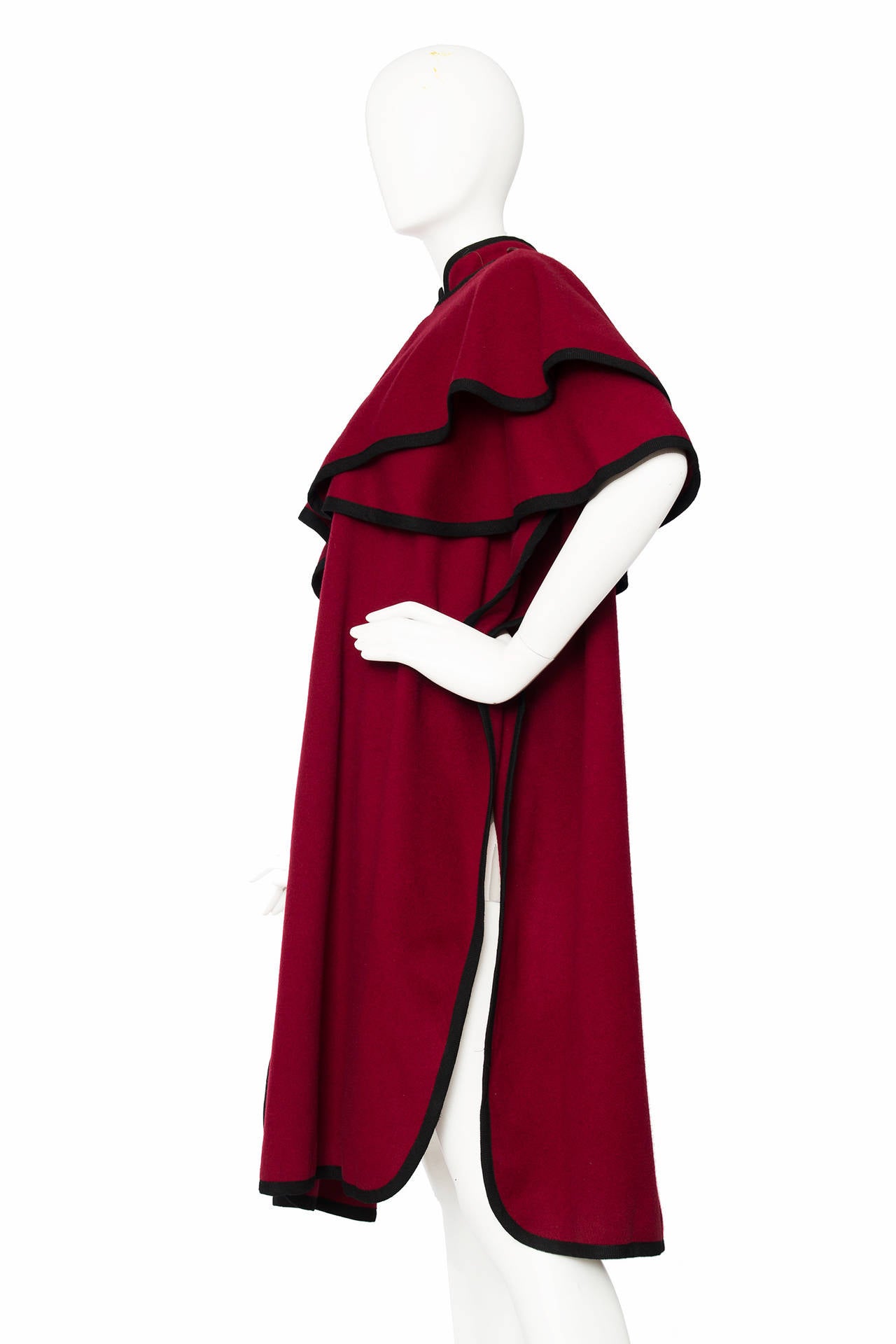 A ca. 1976 Yves Saint Laurent burgundy wool cape with voluminous capelet style sleeves. The cape has a button down front, black trimming and round edges to the hemline, a stand up collar and a single one button closure to the side of the garment.