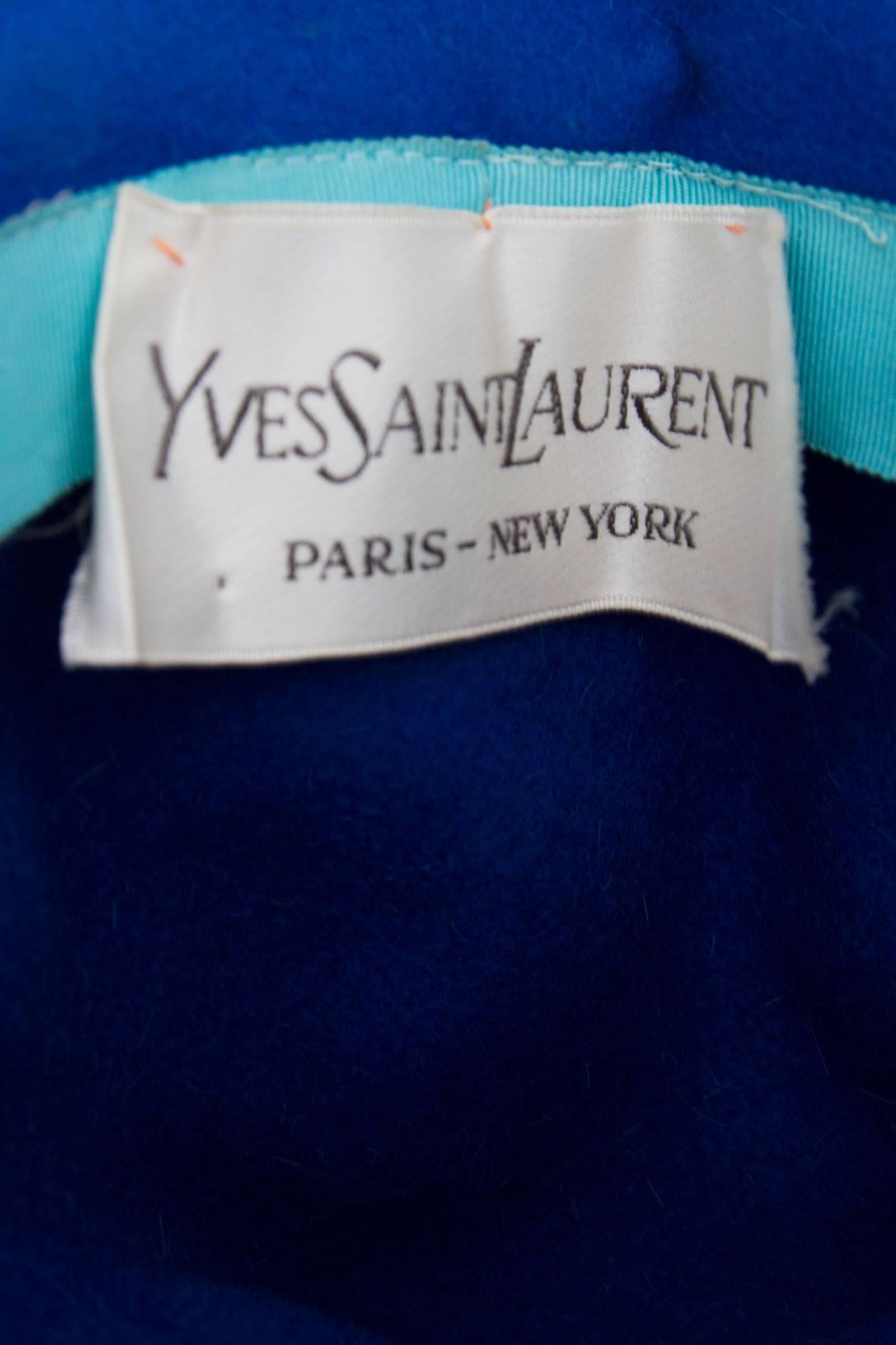 A sculptural 1960s Yves Saint Laurent Blue felt hat with a slim brim and a contrasting orange leather trim. The trim is fastened in each side with a gold button with an anchor detail. On the top the hat is shaped into a handle. 


