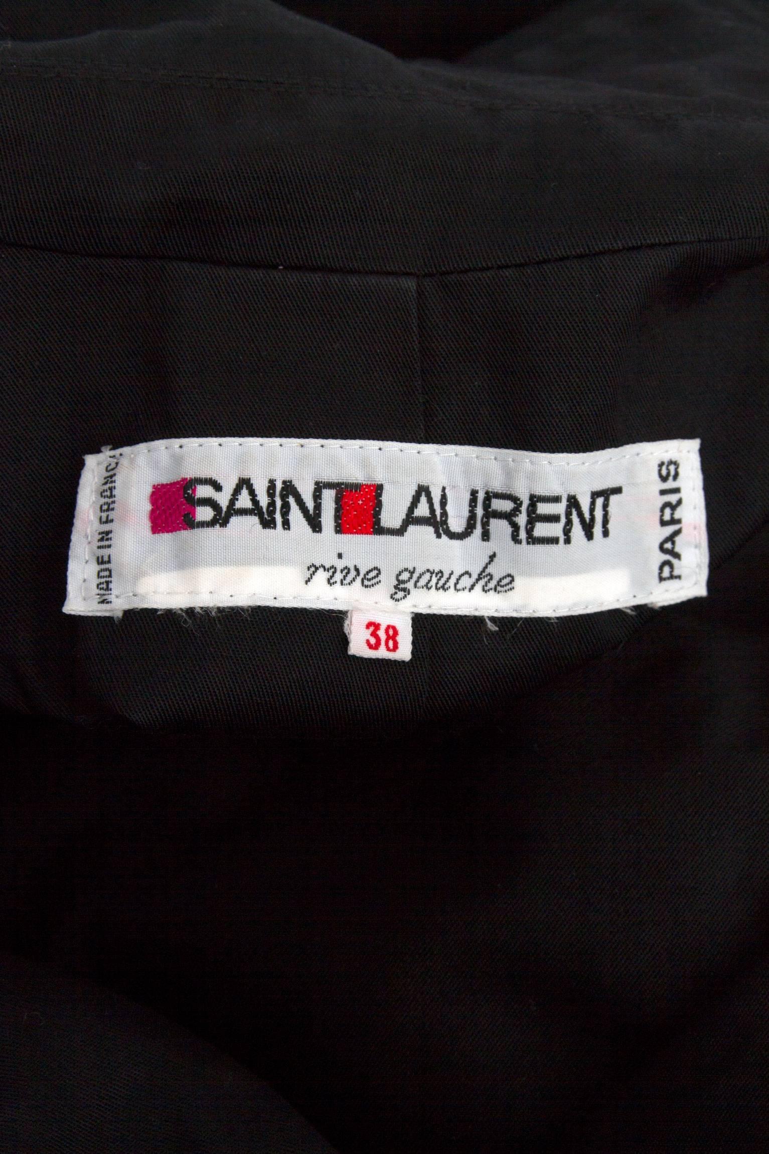 A 1970s Yves Saint Laurent double breasted black cotton trench coat with exaggerated lapels that gently cover the top of the bust and shoulders. A belt around the waist can be tied to define the waistline. The coat has one buttoned cuffs, is fully