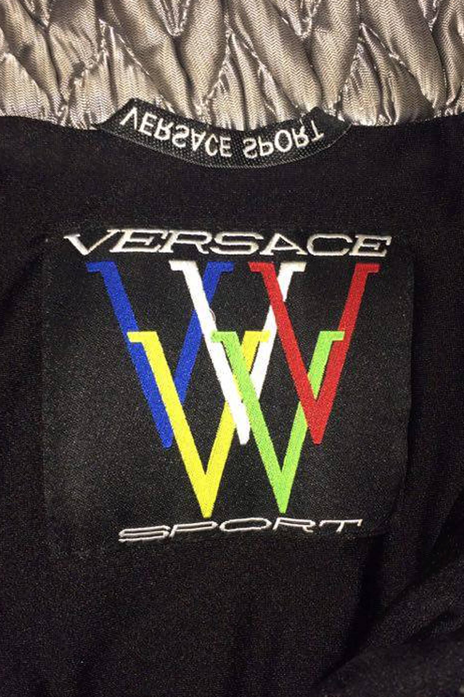 An incredible 1990s Versace Sport one-piece ski suit with black elasticated stirrup pants with tapered ankles and a silver quilted puffer jacket at the top. The detailed jacket has a vertical zipper pocket at the bust and an elasticated clasp belt