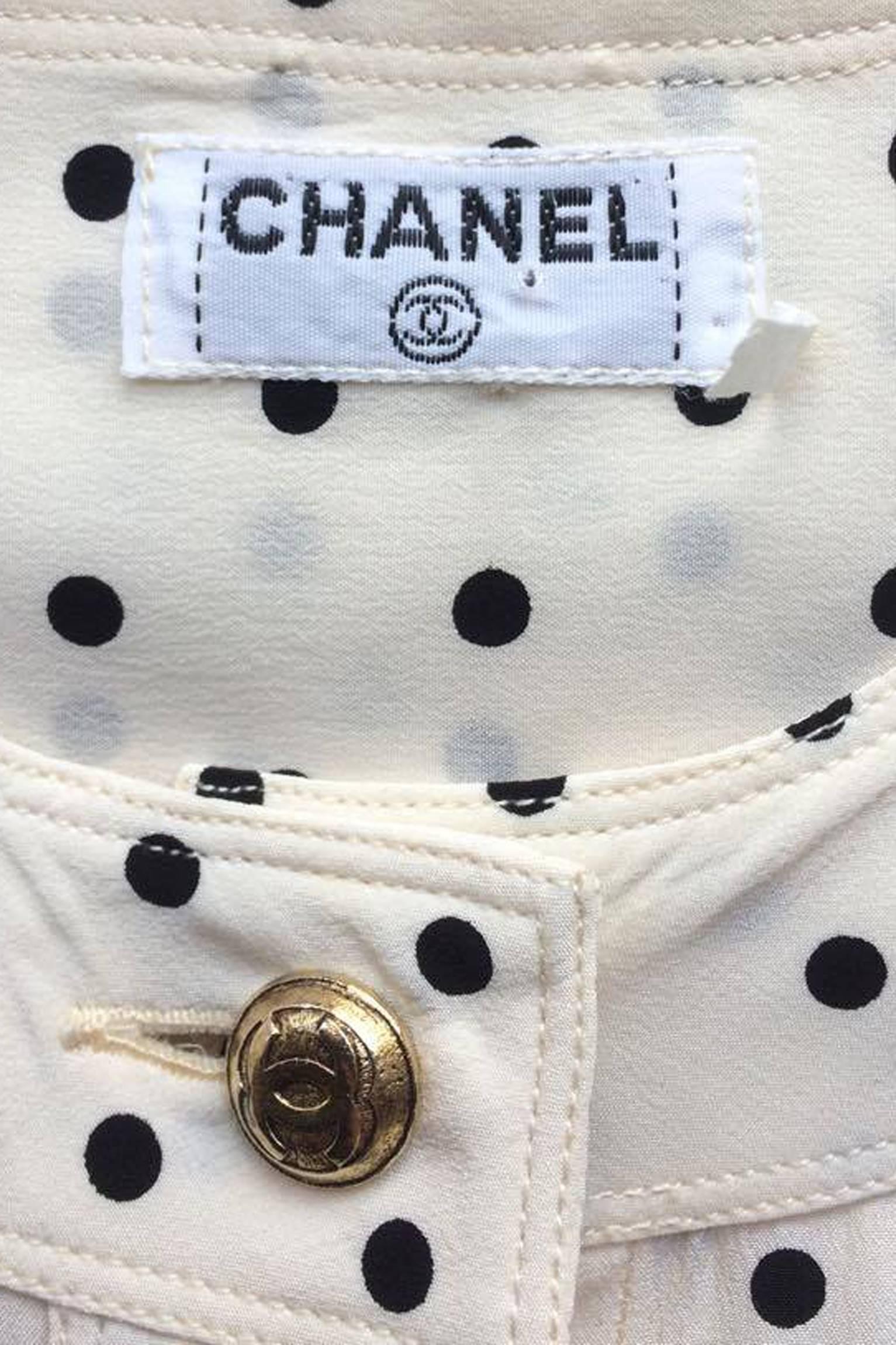 A 1980s white Chanel black polka dot silk blouse with a round neck sans collar, long wrist length sleeves, and a full button-down front. The blouse has a patch pocket situated on each side of the hip just above the hemline. All buttons are gold tone