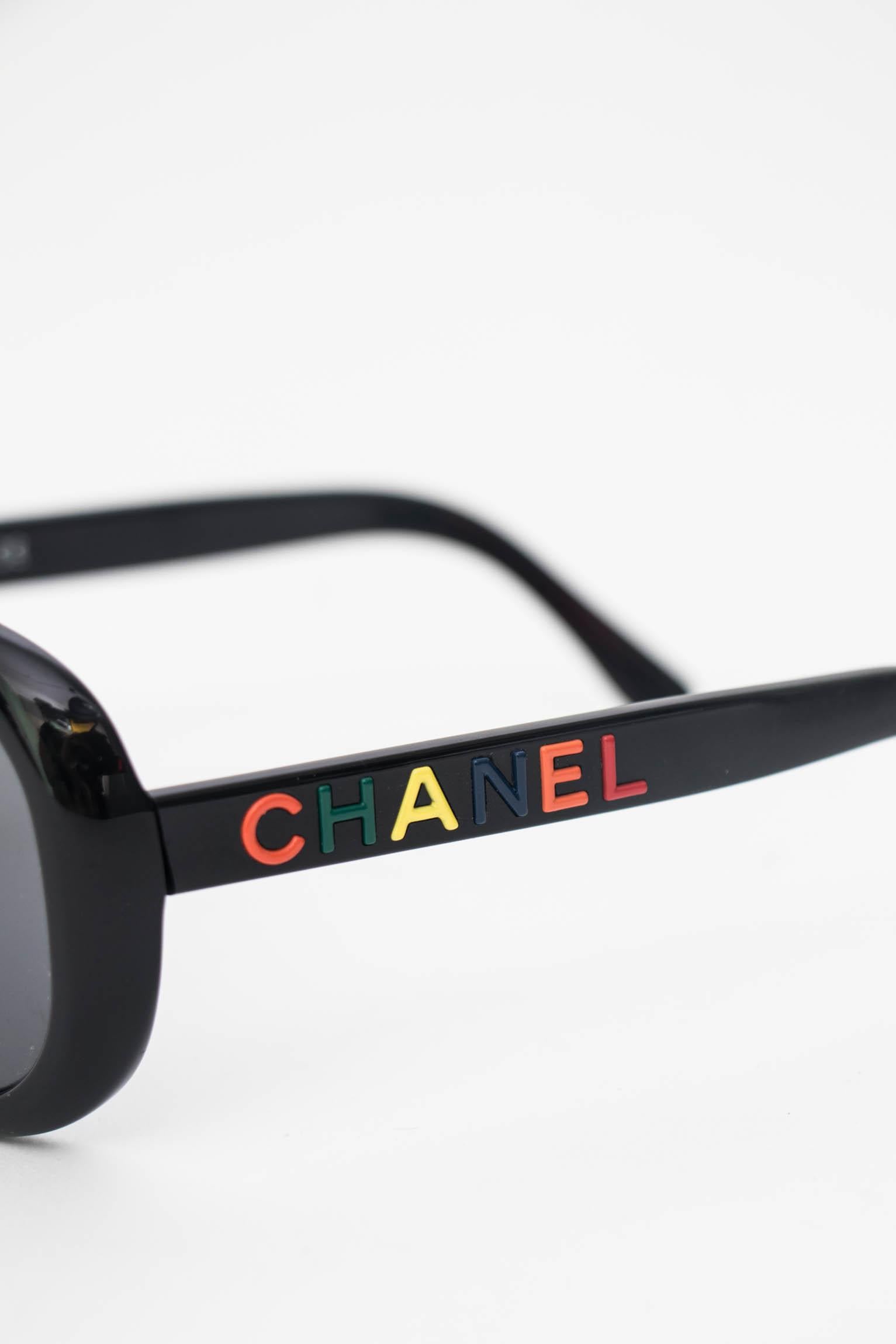 A pair of 1990s black Chanel sunglasses with round glasses and Chanel written down both temples. The letters are held in bold red, green, yellow, orange and blue. 

The sunglasses have the following measurements:
Temple length: 13 cm
Bridge size: 1