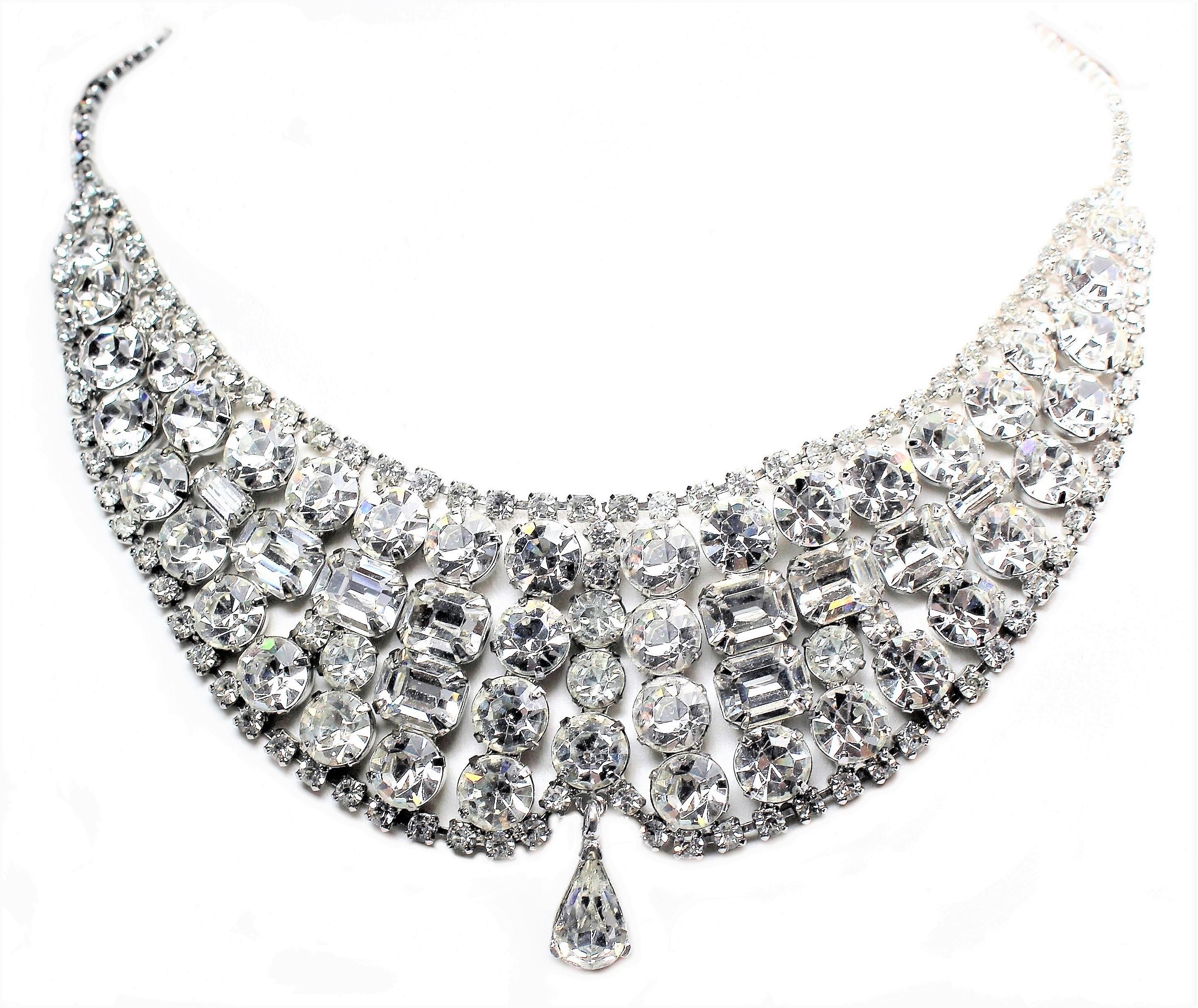 Circa 1950s bib-style cocktail necklace in a bright silver tone metal, prong set with large round and emerald-cut clear faceted crystal stones and small clear rhinestones . It is embellished at the bottom with a faceted pear-shape drop.   The