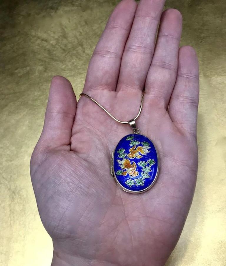 This vintage blue locket is crafted with gilded 925 sterling silver and features a very detailed enamel cloisonné scene, in vibrant shades of blue, green, and orange of Koi fish swimming in a blue lake with green vegetation. The inside of the unique