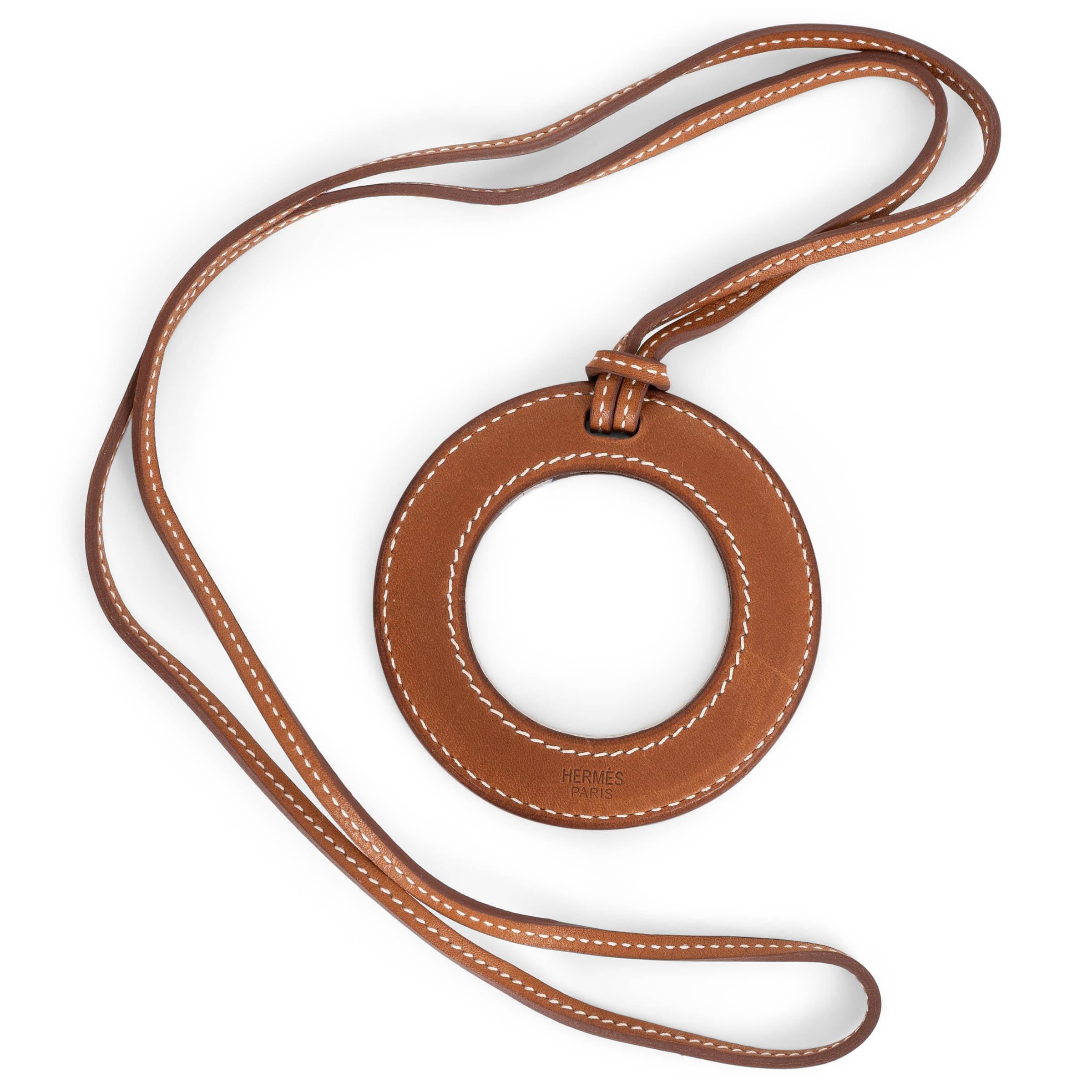 Questa collana Loupe di Hermès, autentica al 100%, è realizzata in pelle Veau Barenia Fauve (cognac) con le tipiche cuciture bianche a contrasto. Caratterizzato da un ciondolo a forma di lente d'ingrandimento incorniciato in pelle liscia, fa parte