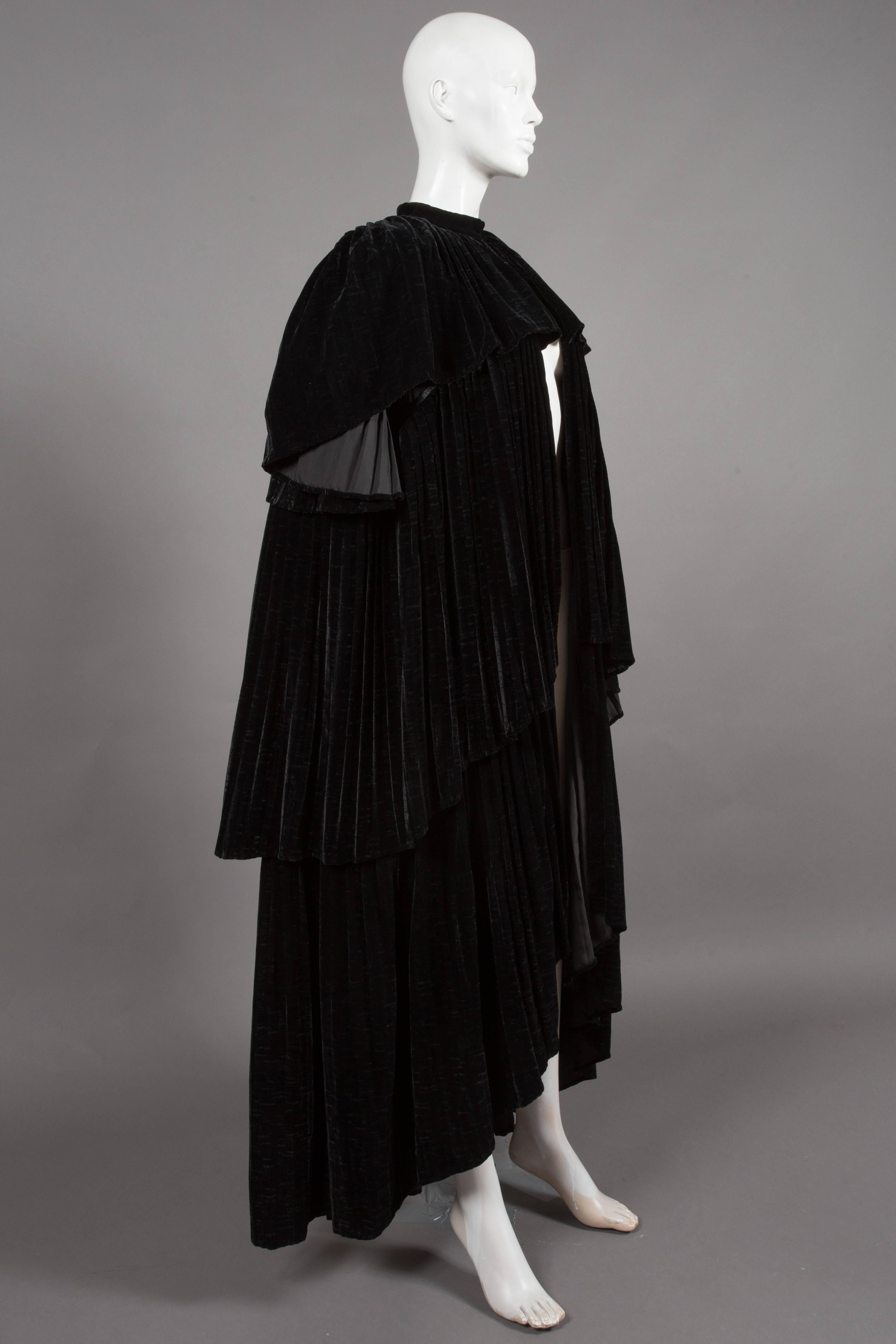 Exquisite Thea Porter Couture black silk-velvet tiered evening cape coat, circa 1970. The cape features heavy pleats through out, three tiers and hook-and-eye closures on collar. The cape is shorter at the front and gradually gets longer towards the