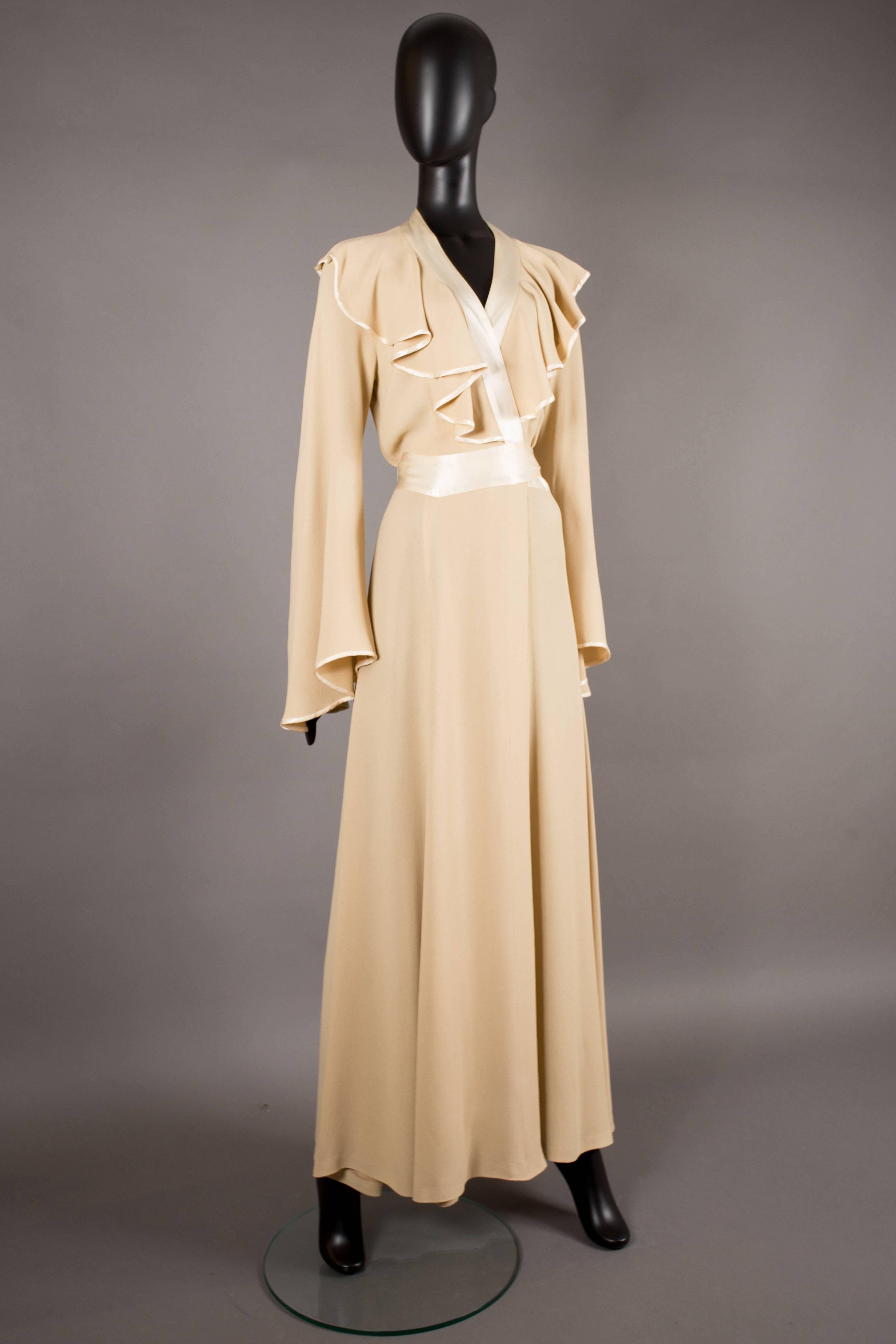 An Ossie Clark evening wrap dress, circa 1970. Champagne moss crepe, ivory satin trim, sash fastening at the rear, angel sleeves and ruffled collar. 

Size: 
Shoulder to shoulder - 18