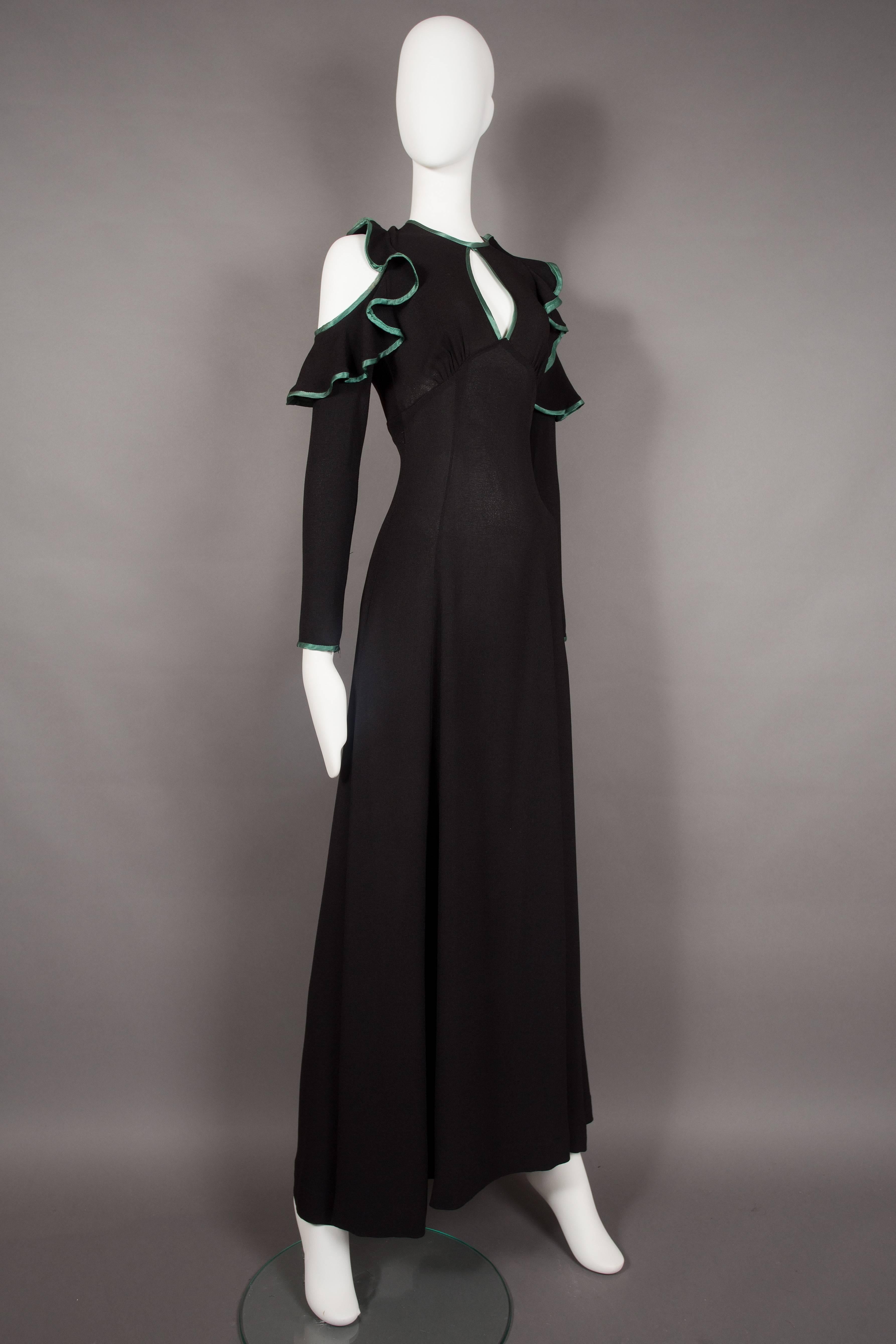 An Ossie Clark black moss crepe evening dress, circa 1968. Cold shoulder design with ruffles, green satin trim throughout, peek-a-boo cleavage, open back, bow fastening and metal zip closure on waist. 