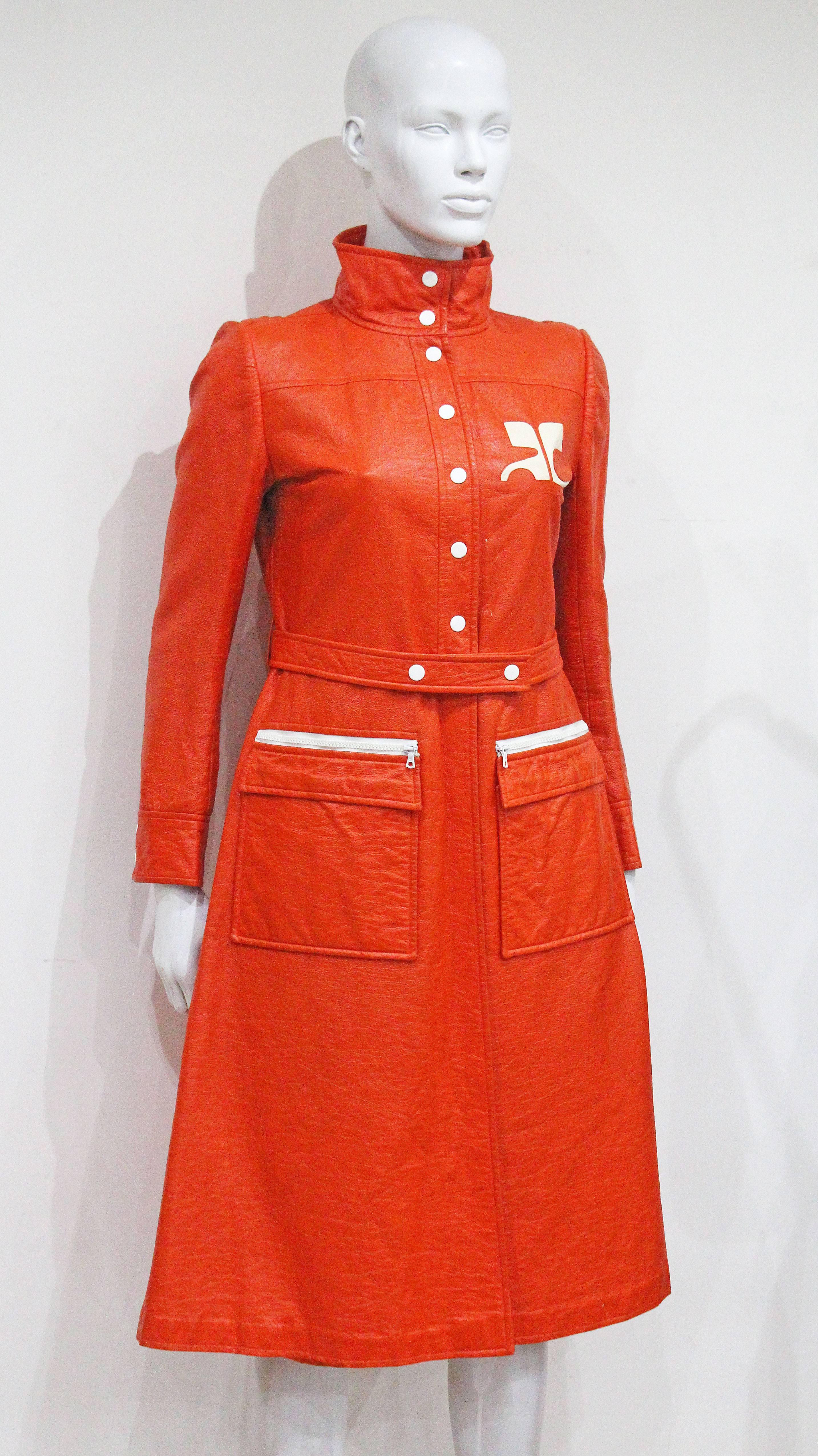 A Courreges coat dress from the year 1970 in orange vinyl fabric. The coat dress features stand collar, matching belt; white dot-button at centre-front and large Courreges logo on left breast. 

S/M