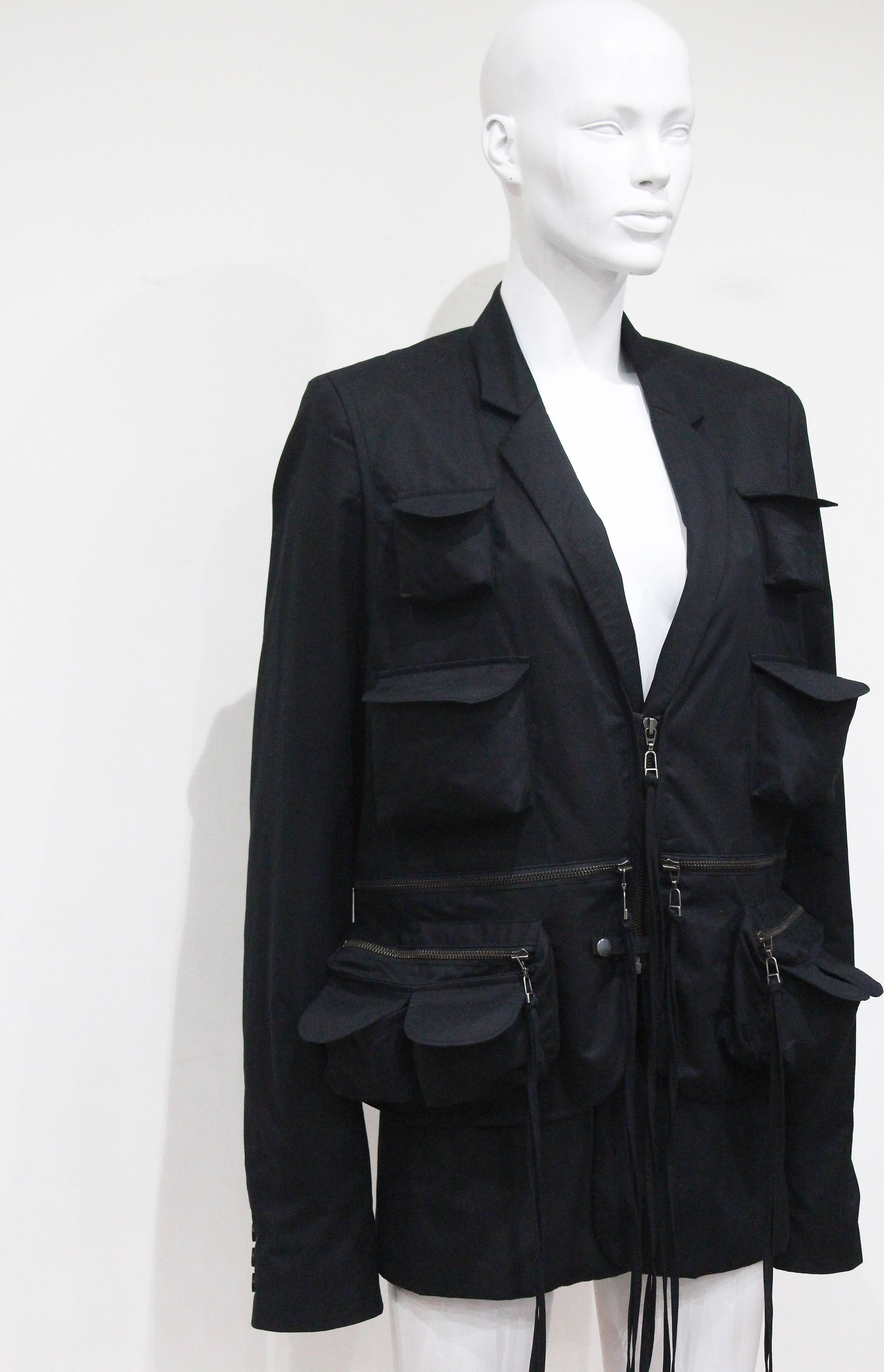 A Raf Simons black military style blazer jacket from the spring/summer 2003 'Consumed' collection. The blazer jacket of black cotton has an attached military waistcoat with multiple pockets with extra long tasseled zippers. 

Mens Size 50 