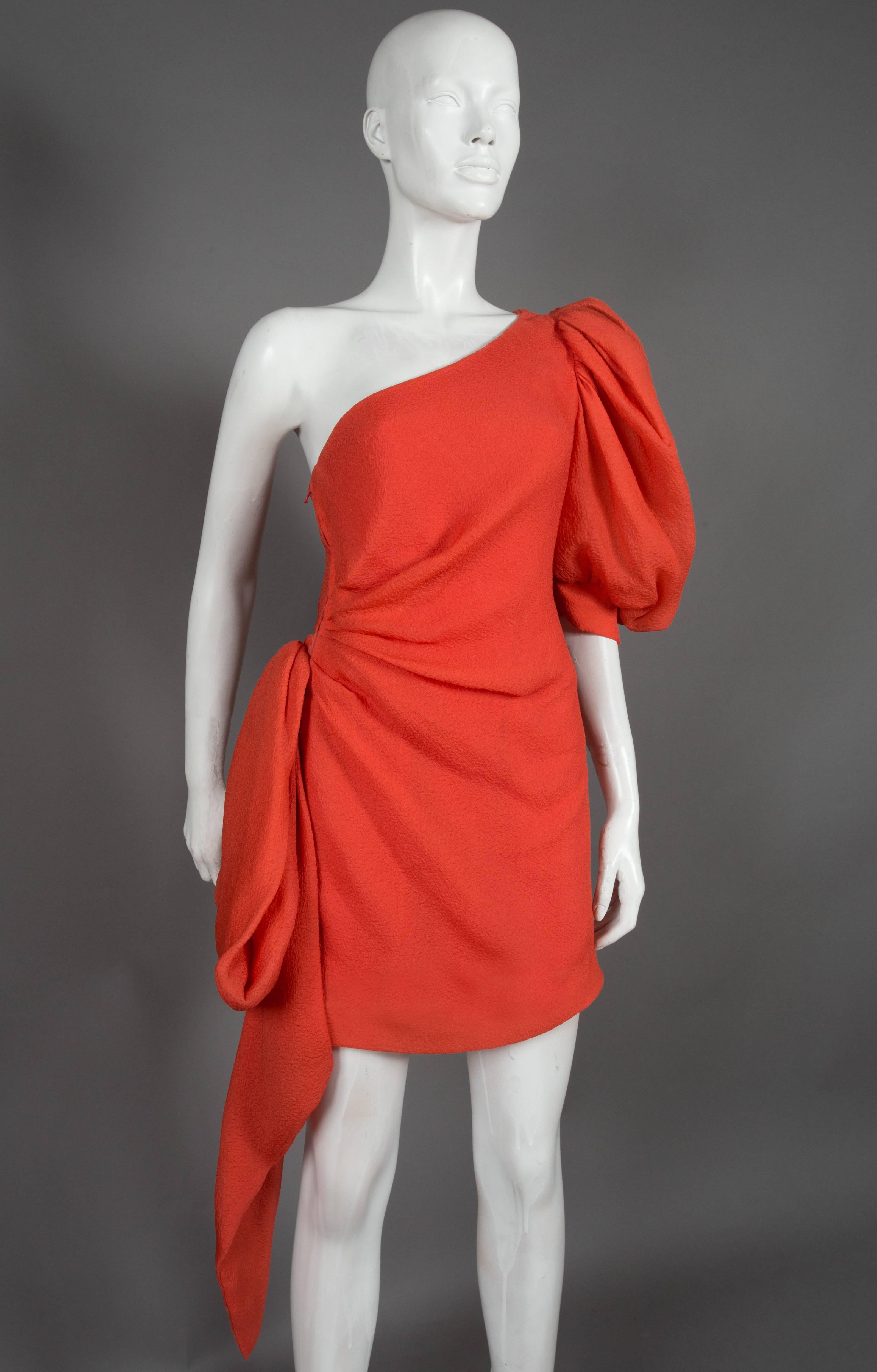 This is a beautiful asymmetric cocktail dress designed by the great Hubert de Givenchy, the dress is in his signature coral colour in a 100% silk crêpe and features a balloon sleeve and an extravagant bow which folds to the front. 

This dress