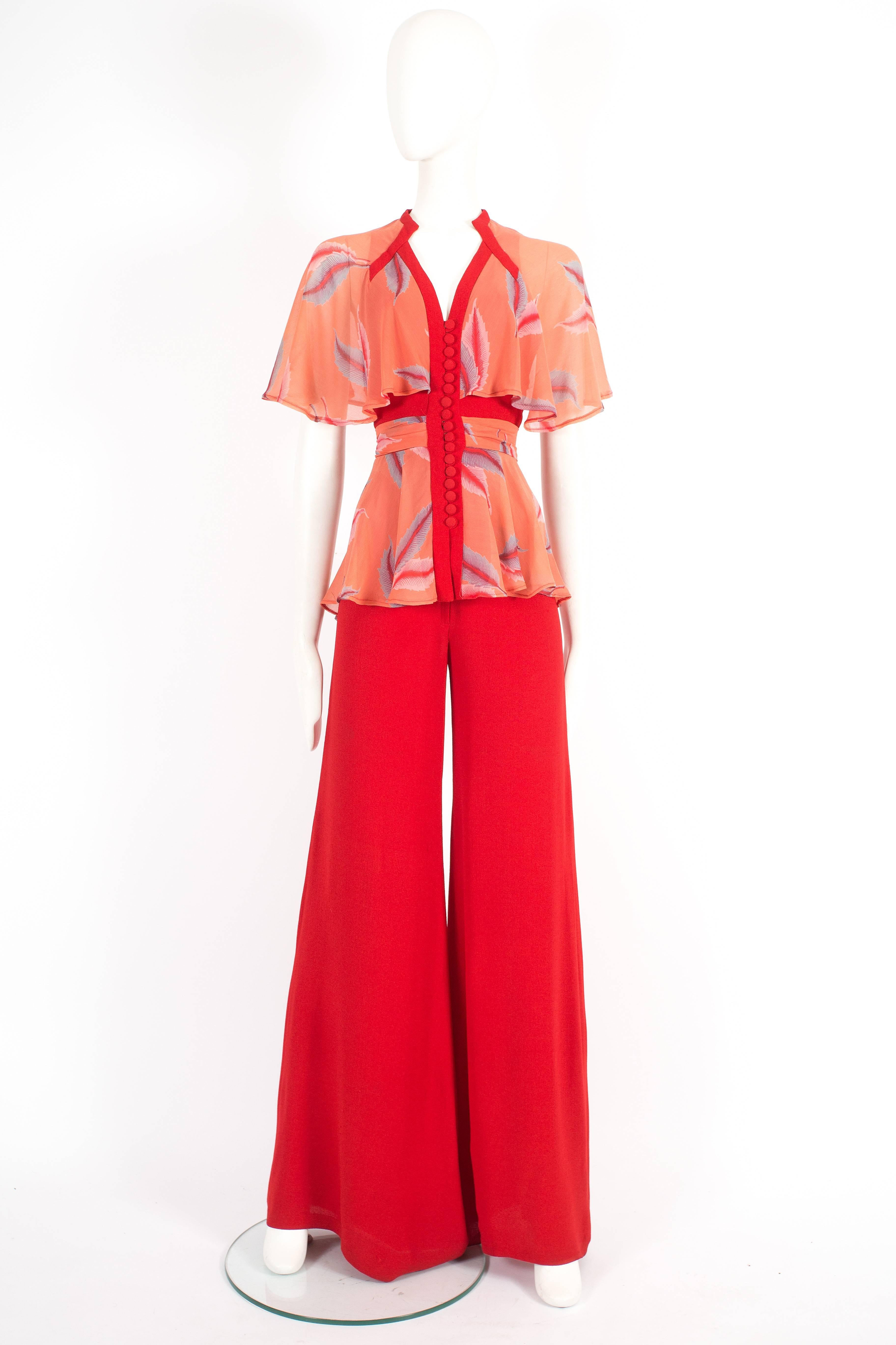 Xenia pantsuit, circa 1970. Constructed in red moss crepe and chiffon, features button fastenings, waist tie, high waisted wide leg pants and capelet. Very Ossie Clark!

