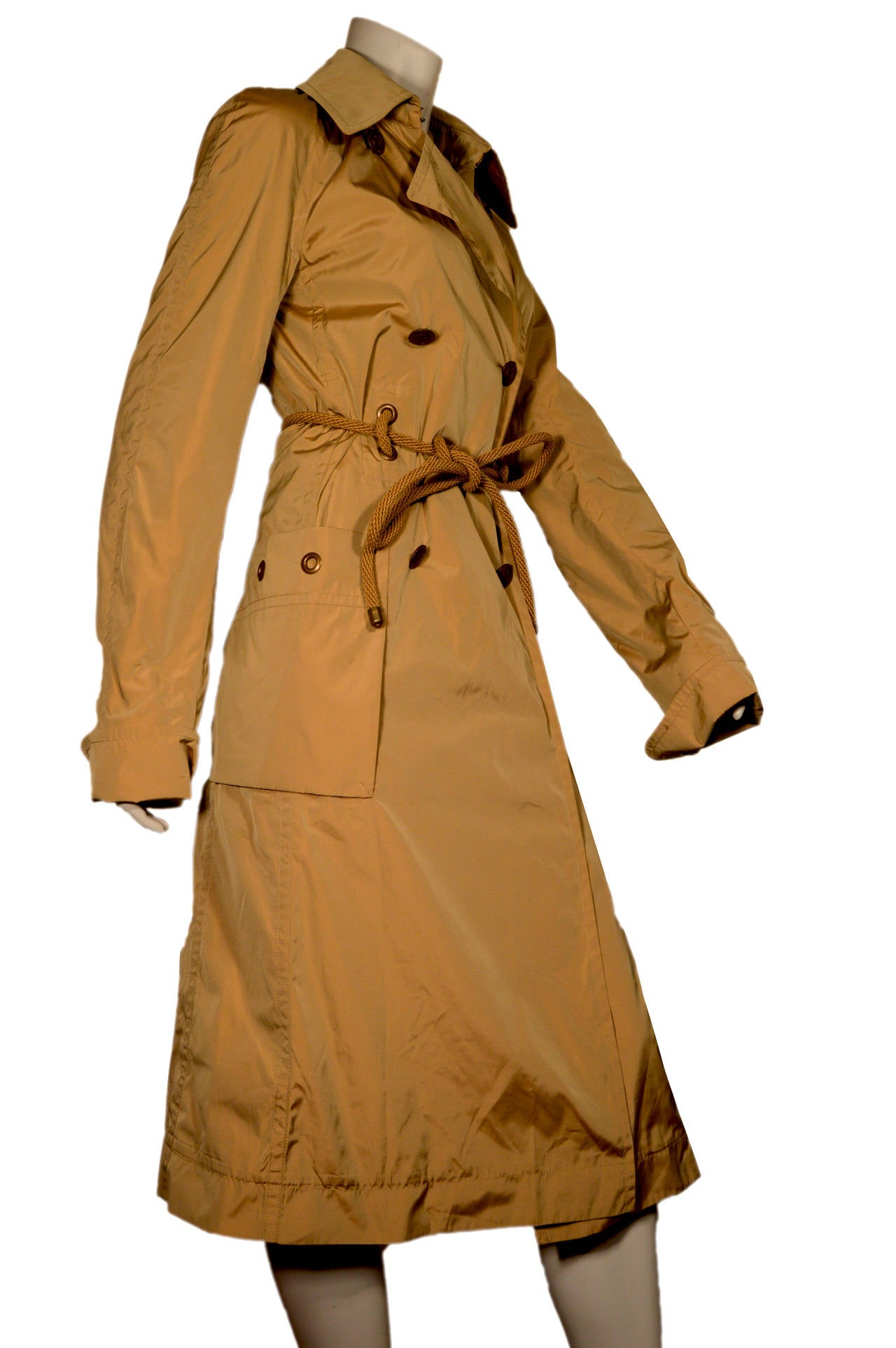 This double-breasted trench coat from YSL is an iridescent beige. There are metal buttons down the front, as well as metal grommets featured decoratively on the pockets, cuffs and belt. There are two oversized pockets on the front. The coat is