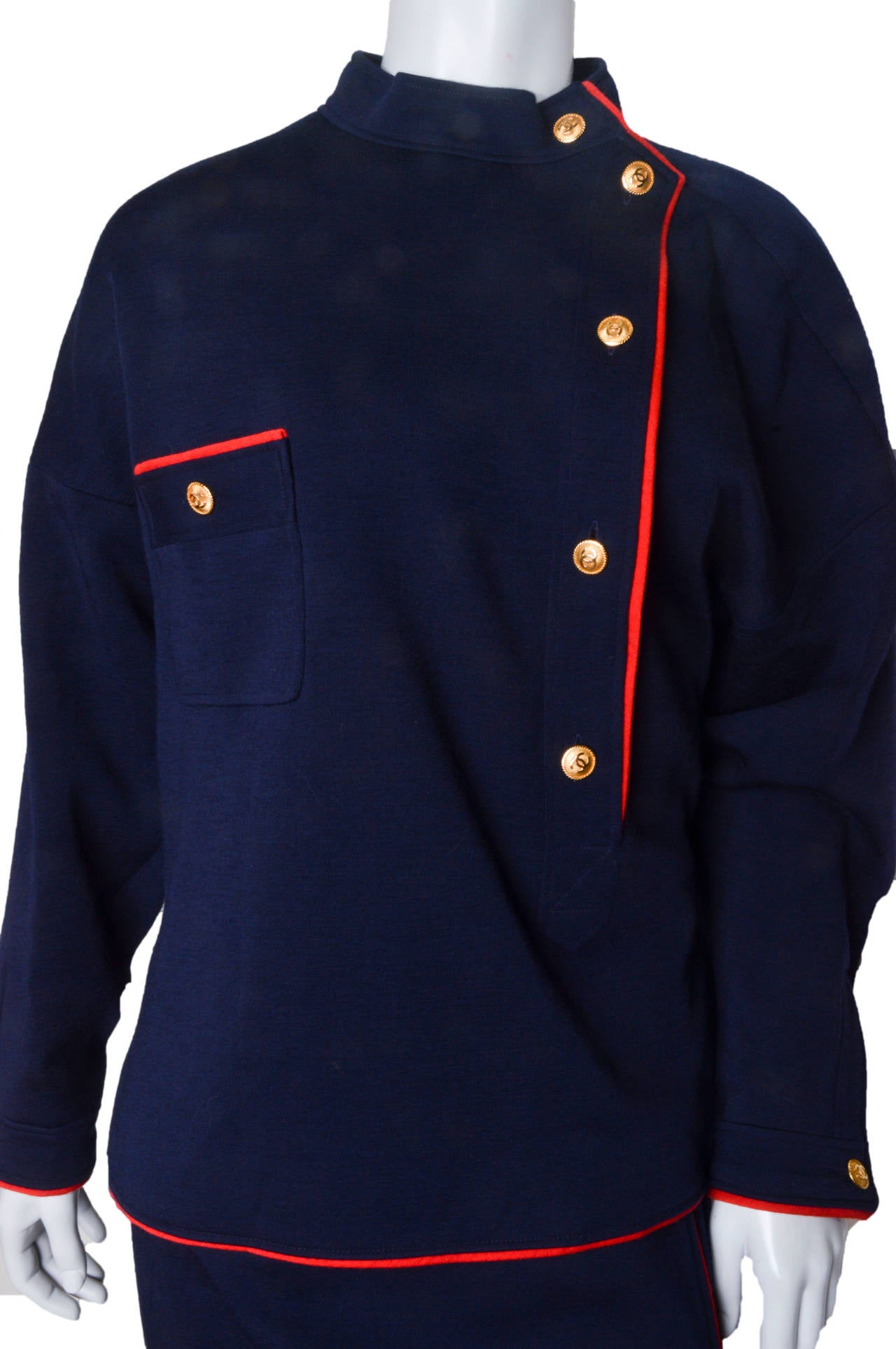 Fabulous vintage Chanel Boutique knit Suit.
Style number as tagged: 20153.
Very military inspired navy blue with red piping.
Pull over top features asymmetrical gold Cc embossed buttons 
with red piping.
High neck. Slit pocket on chest with