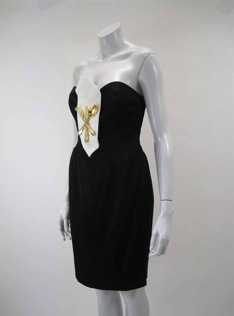Rare and iconic Moschino Dinner dress.

From his 1989 Fall "Show Off" Couture collection.

Black strapless with folded white embroidered napkin with gold tone metal knife, fork and spoon.

Side zipper.

Fully lined.

Tagged size US