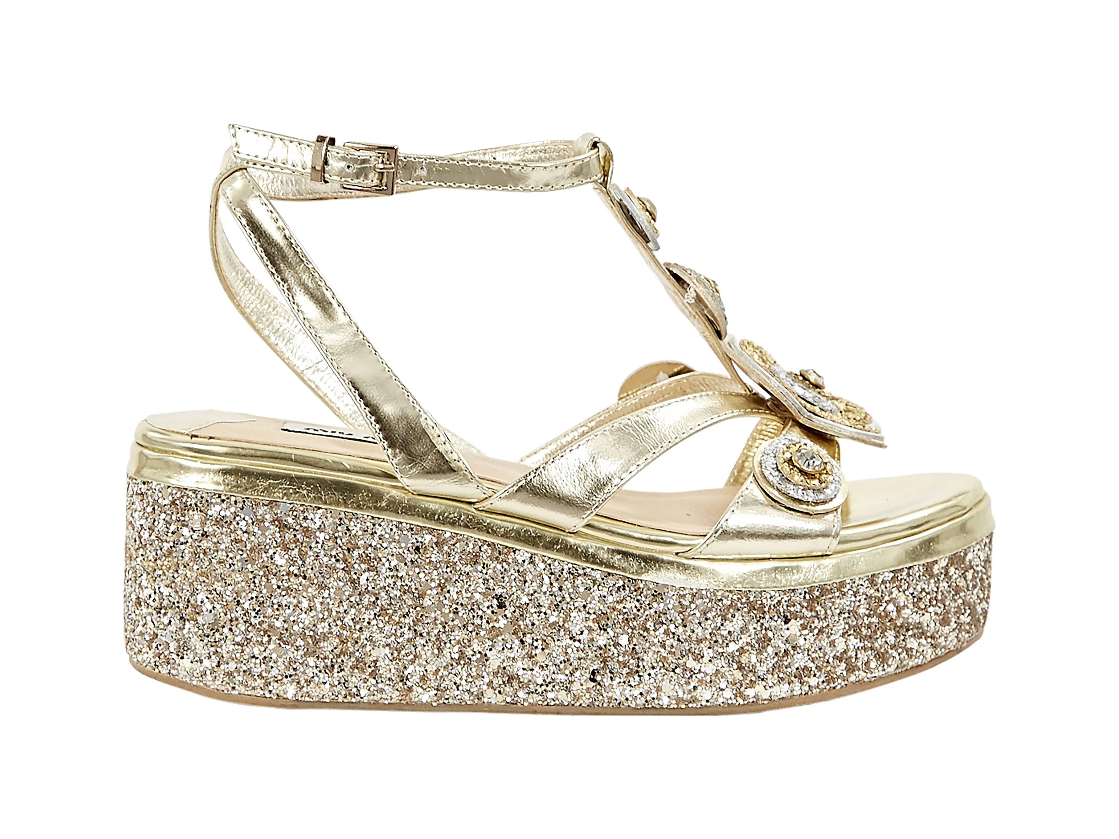 Gold leather and glittered wedge sandals by Miu Miu.  Adjustable ankle strap.  T-strap accented with embellished floral and round details.  Open toe.  Glittered platform wedge. 