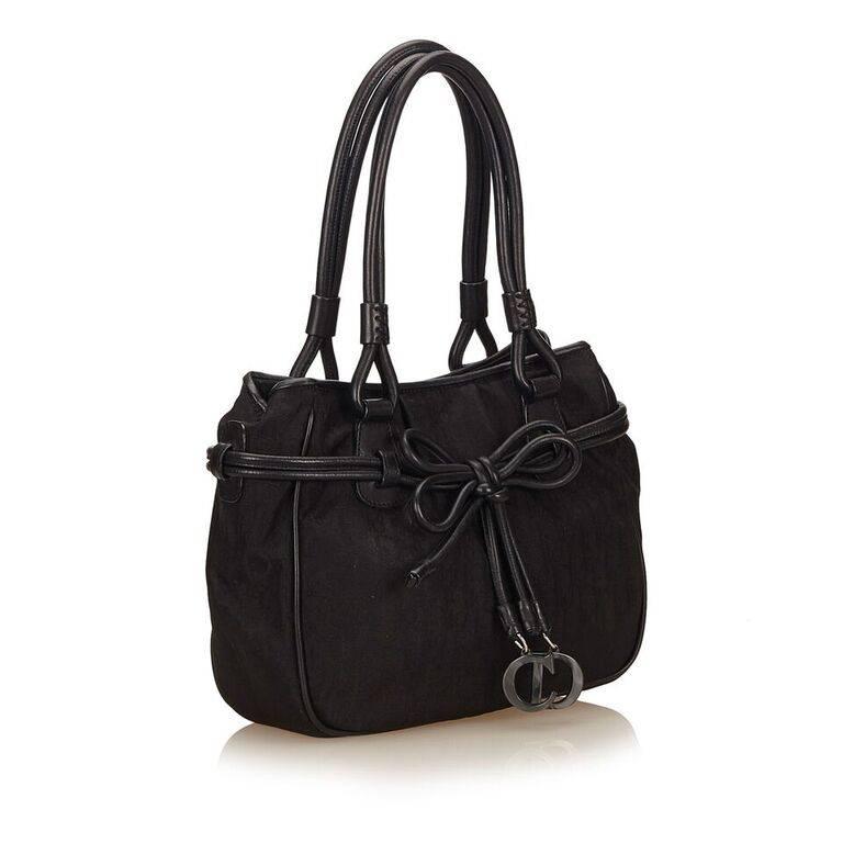 Product details:  Black nylon shoulder bag by Christian Dior.  Trimmed with leather.  Dual shoulder straps.  Magnetic snap closure.  Bow with hardware logo charms accent front.  Lined interior with inner zip and slide pockets.  9.5