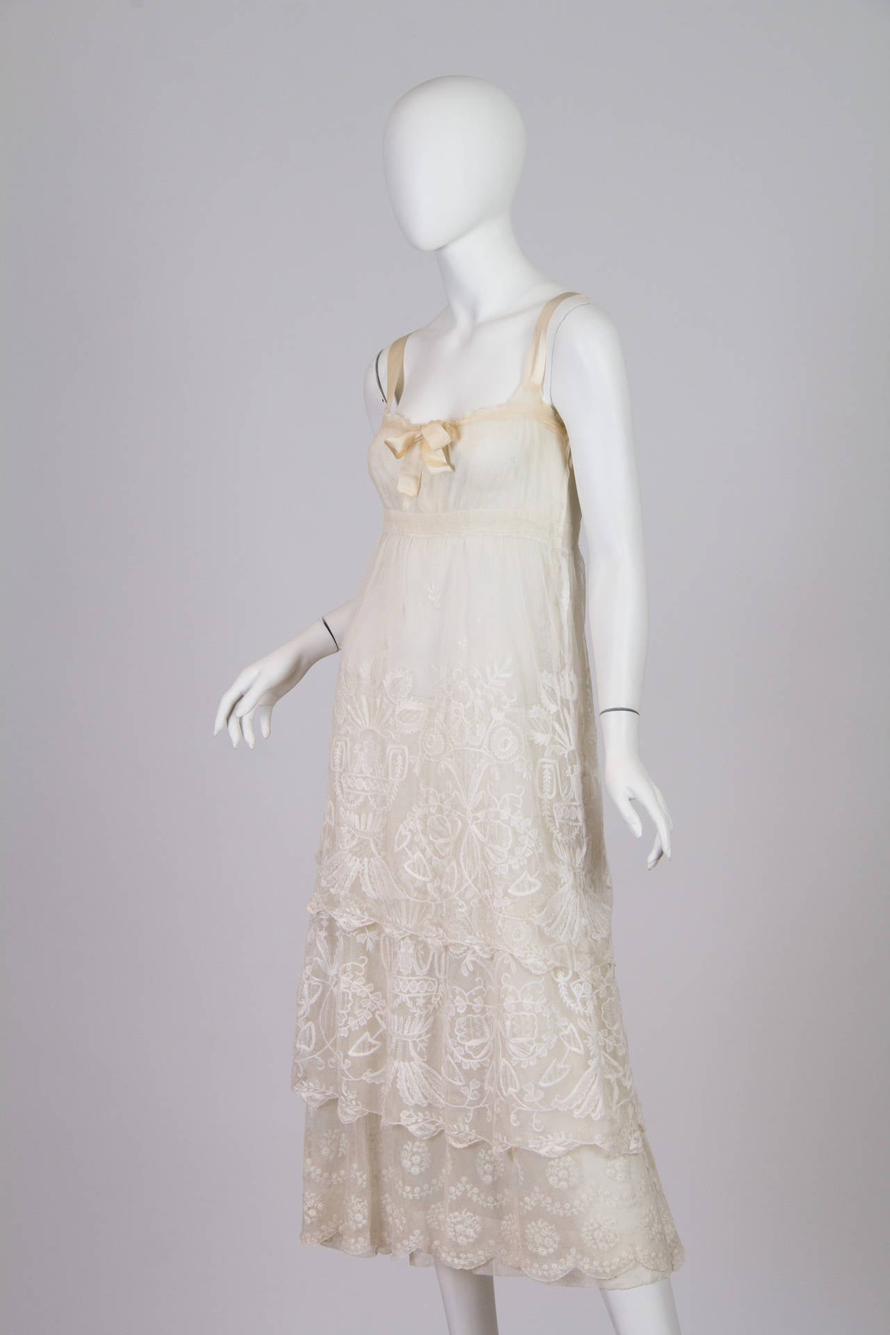 This is a lovely dress made in the later years of the 1910s, as Europe approached World War I. It is sheer cream cotton with an empire waistline and peach ribbon accents at the neckline and shoulders. The stunning white princess embroidery falls in
