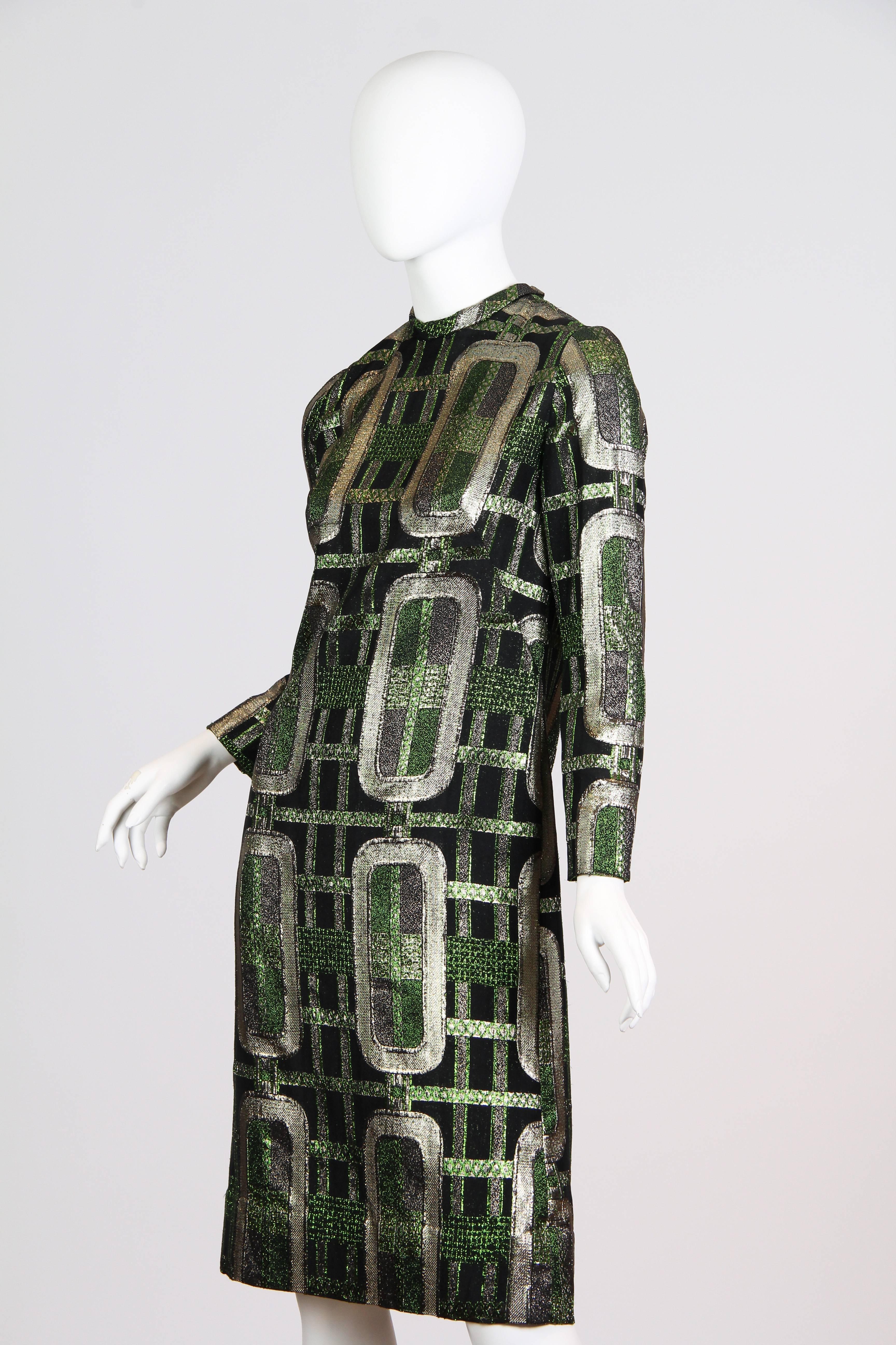 This is a sheath dress of metallic silver, green, and black brocade. Likely dating from the 1960s, the dress is made of a luxurious textured weave in shimmering metallic fibers. The geometric pattern is made of a grid overlaid with chain-like silver