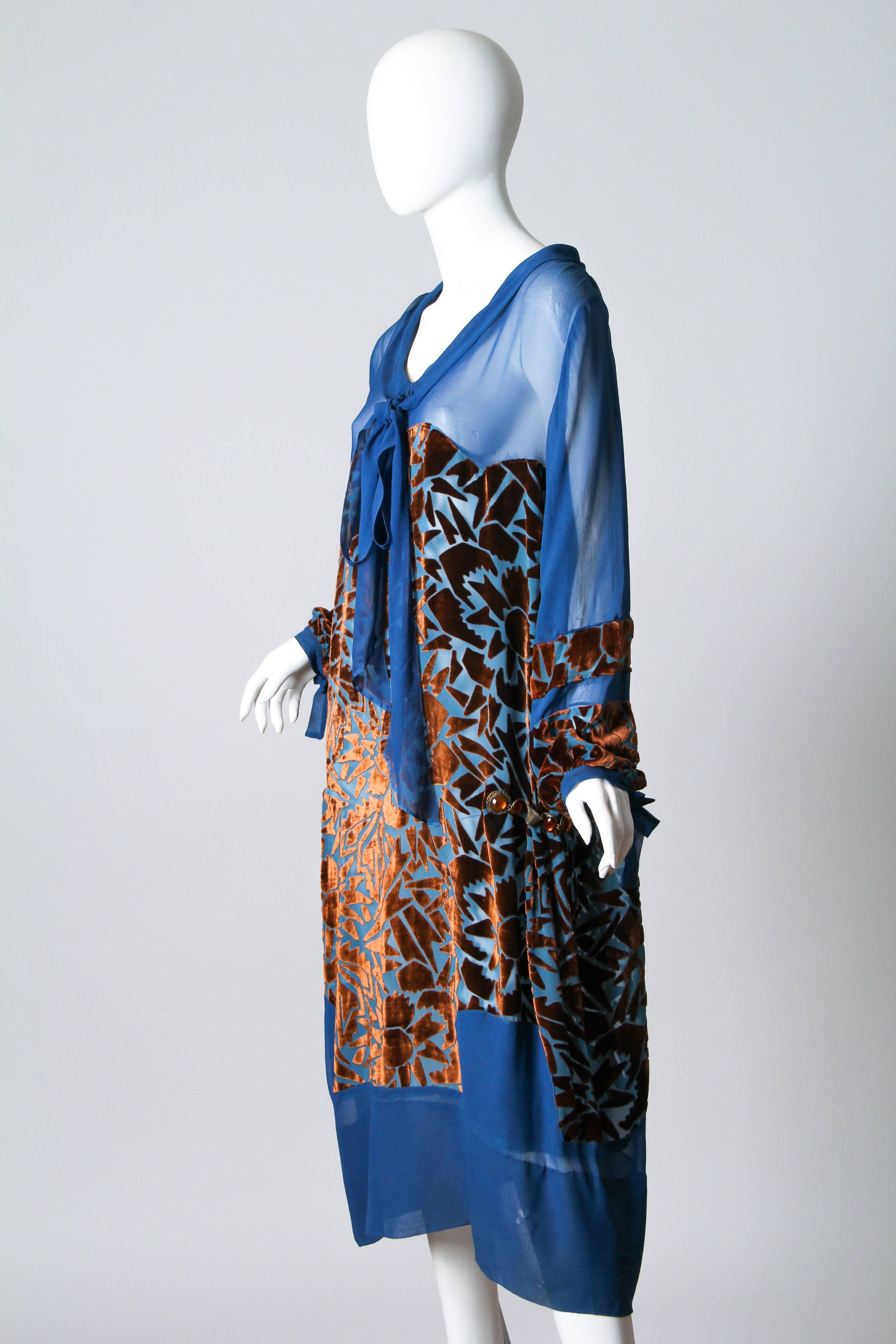 This is a lovely blue silk afternoon dress from the mid-1920s. The sheer base fabric is embellished from the bust to the knees and around the lower arms with beautiful chestnut-brown voided velvet. The velvet is cut in an abstract geometric pattern