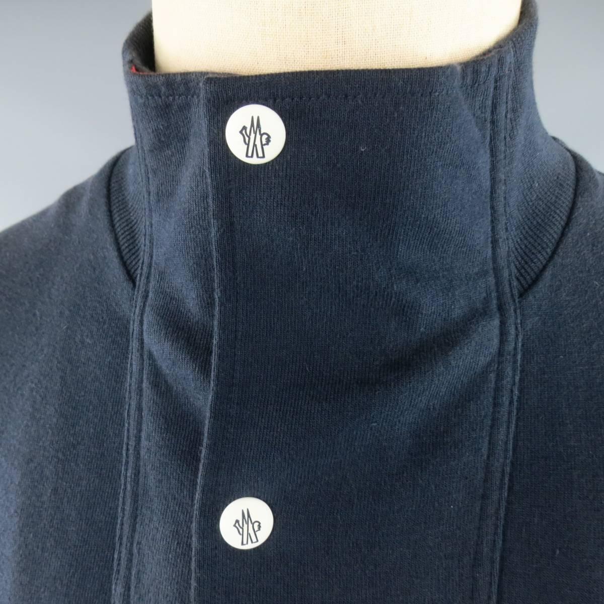 MONCLER GAMME BLEU jacket in a navy blue cotton jersey featuring double zip pockets with white logo tabs, zip up front, and high collar with snap placket and red lining. Fading and wear throughout and discoloration on collar. As-is. Made in Italy.
